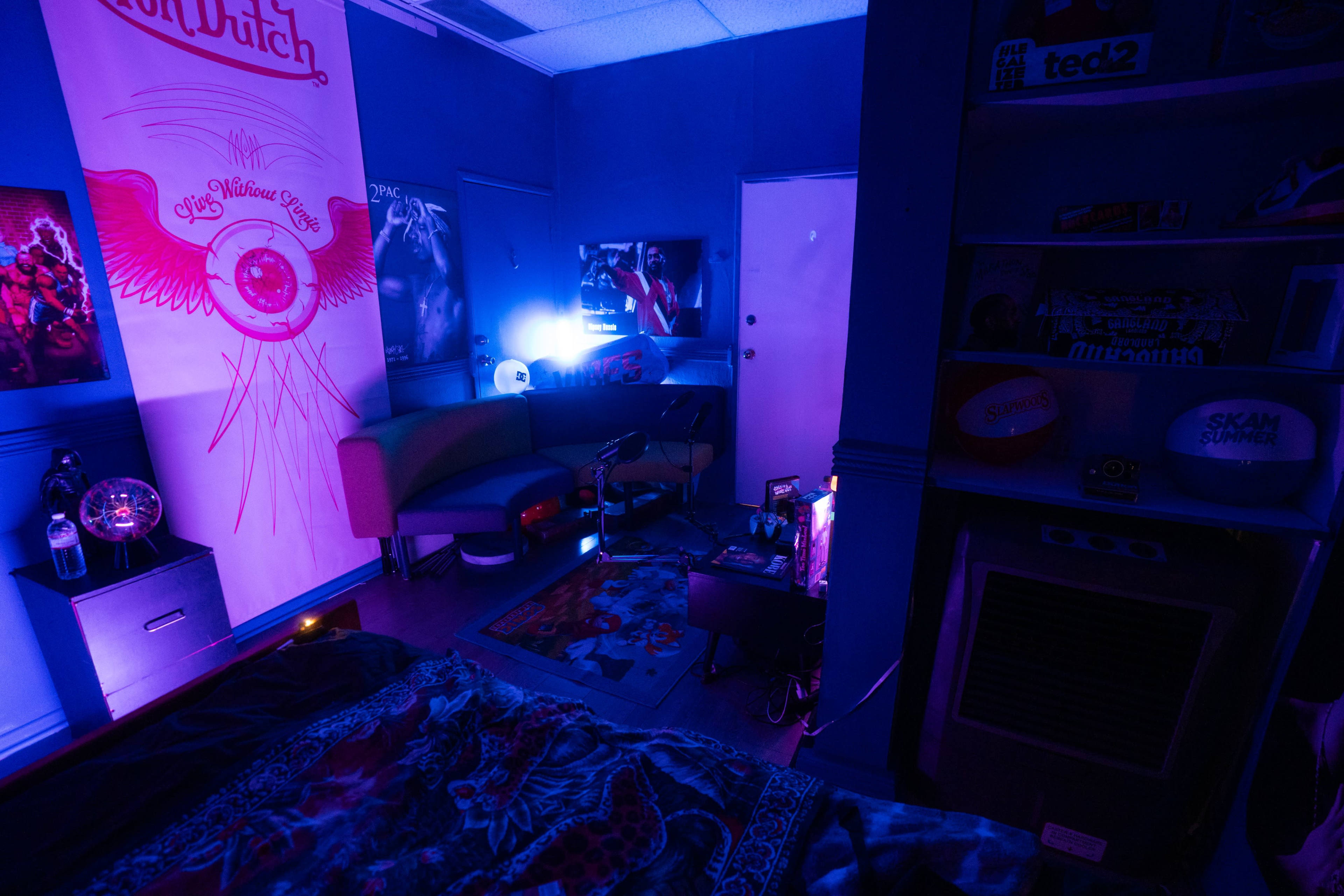 The image shows a dimly lit room with blue walls, featuring a couch, a rug, and various posters and items neatly arranged around a small entertainment area.