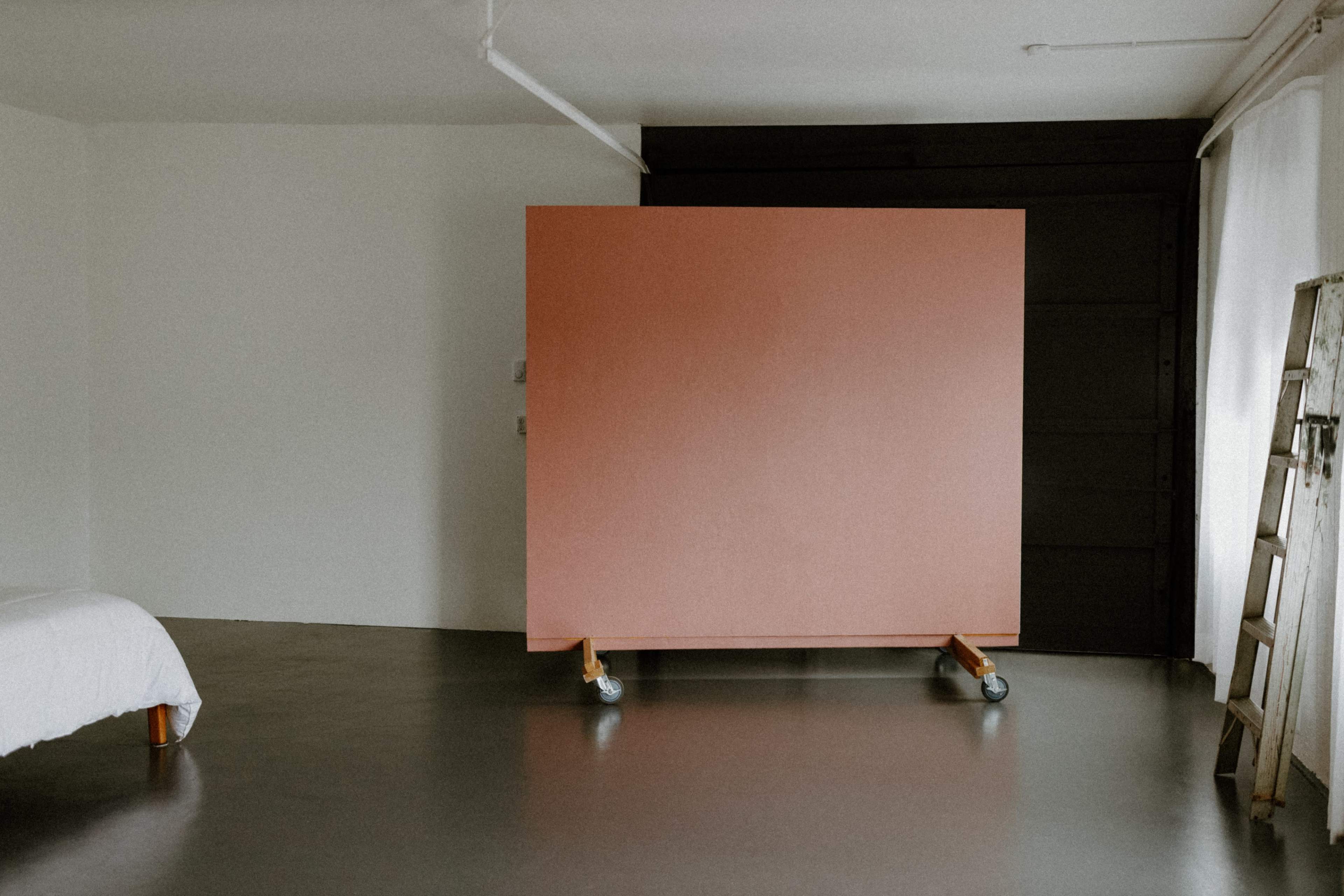A large pink structure on wheels stands in an empty room with a bed and a ladder.