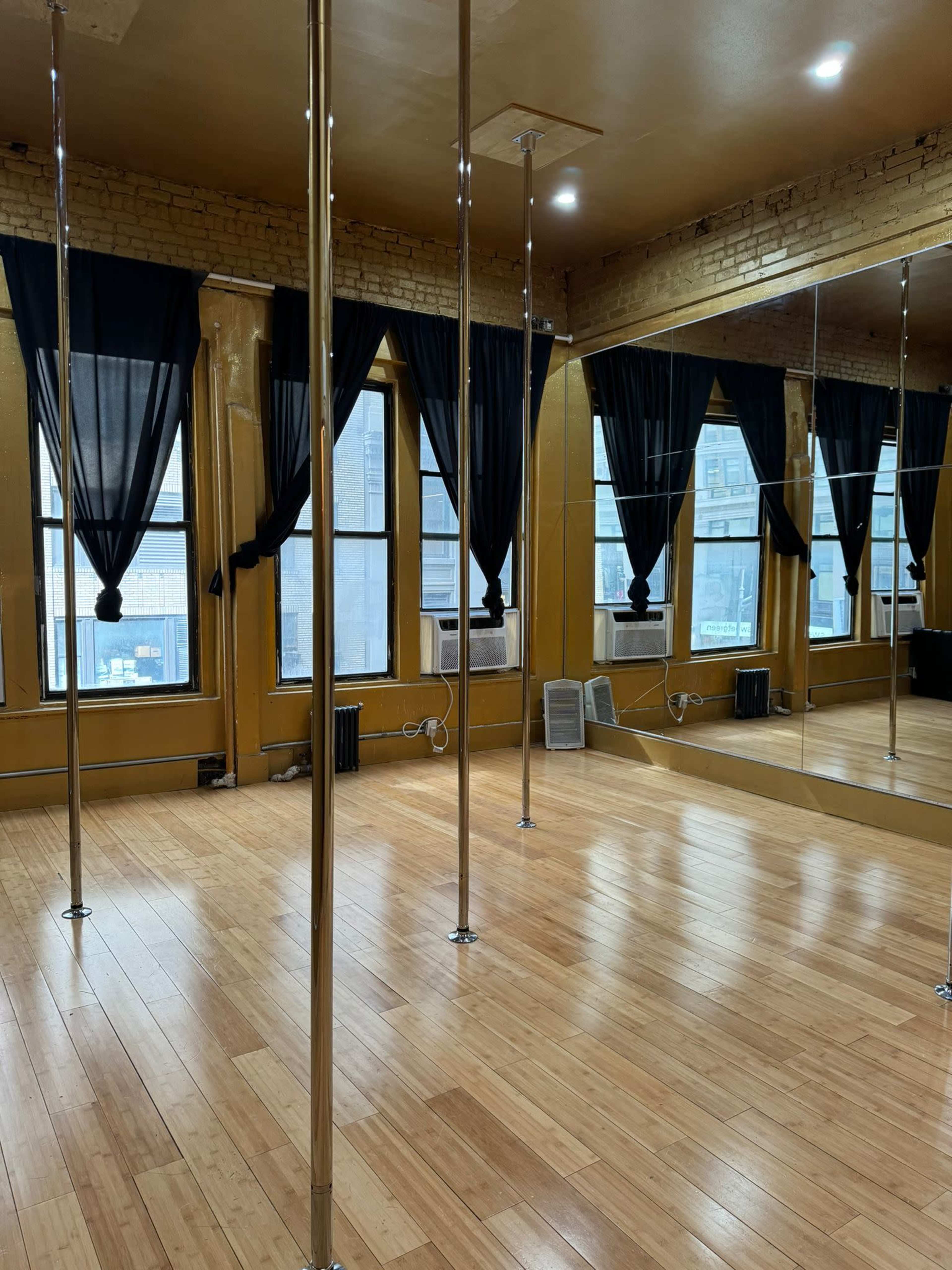 A dance studio with wooden floors, several polished metal poles, large windows draped with black curtains, and a mirrored wall.