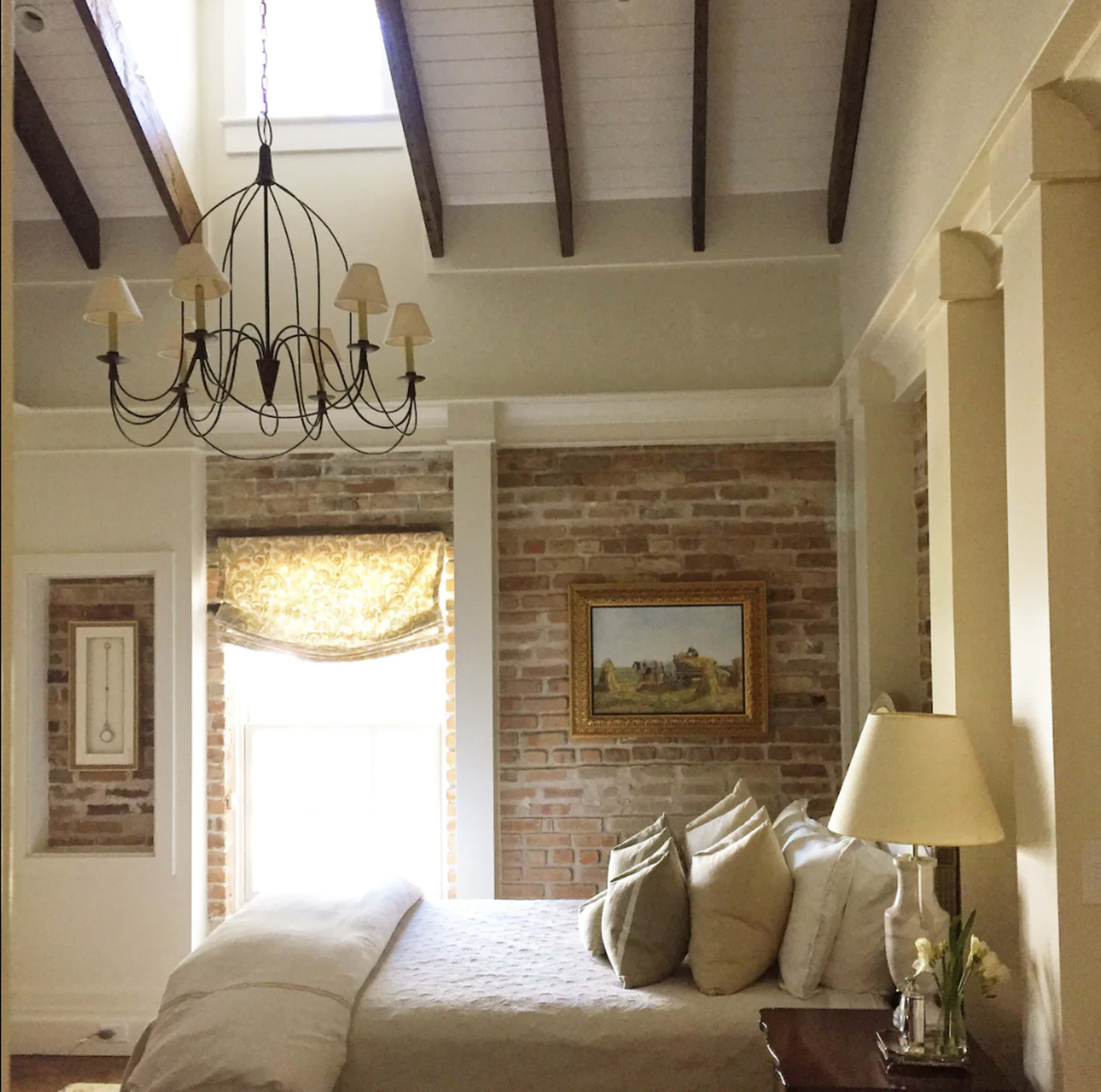 A bedroom featuring a bed with decorative pillows, a chandelier hanging from the ceiling, and a window with a patterned valance, against a backdrop of exposed brick walls.