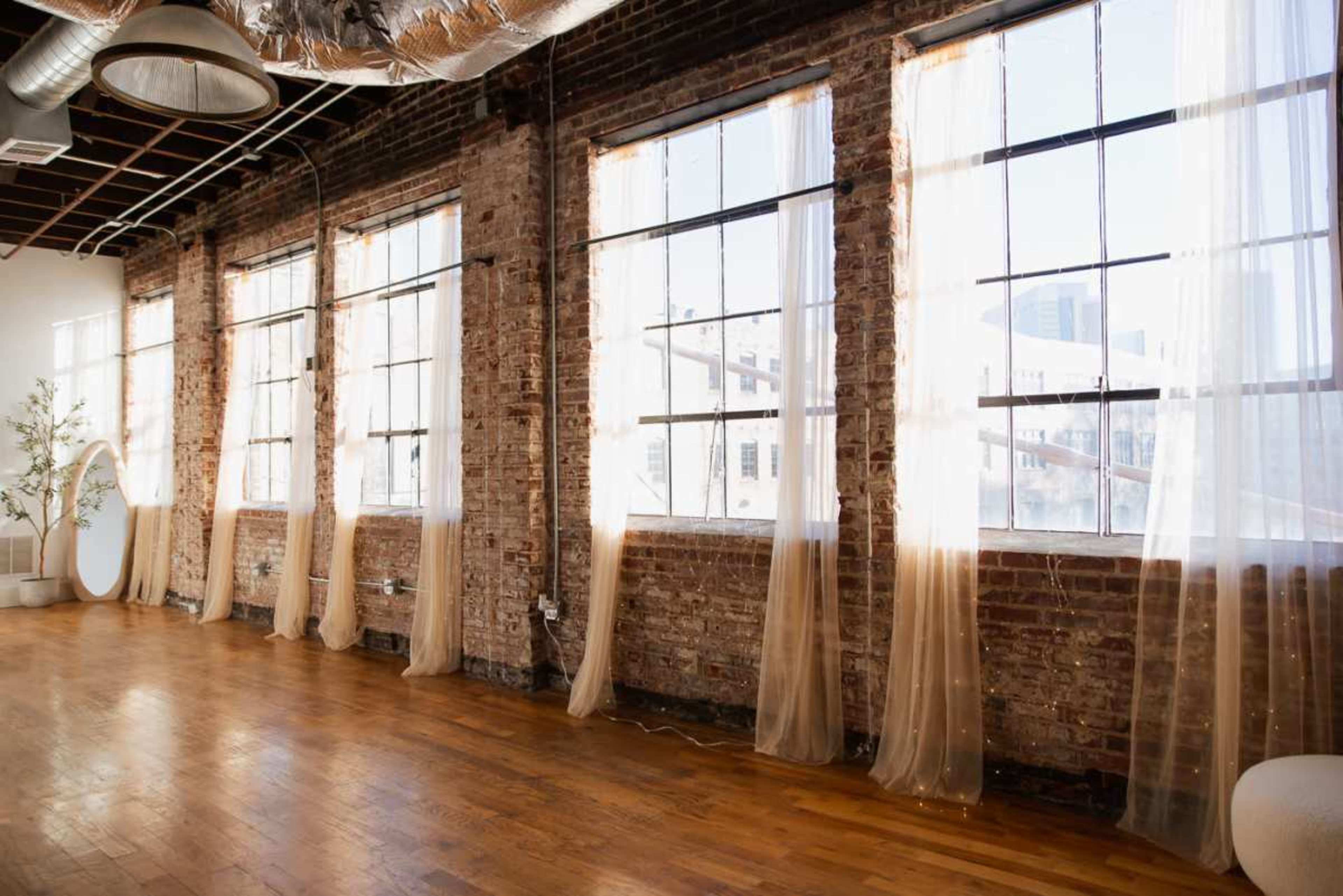 The image shows a spacious room with large windows featuring sheer curtains, exposed brick walls, and wooden flooring.