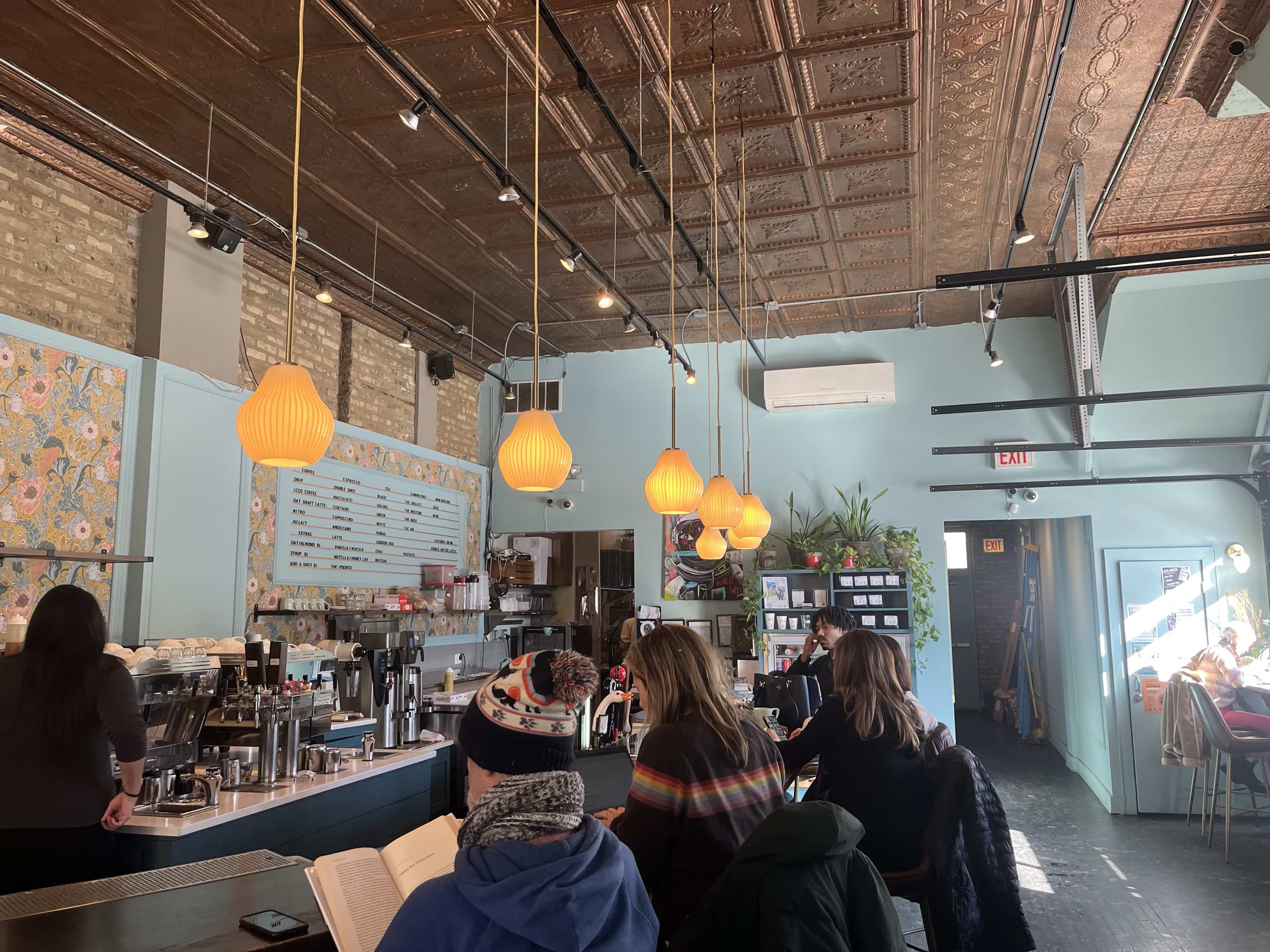 The image shows a cozy café interior with hanging orange pendant lights, a colorful floral accent wall, and patrons seated at tables while engaged in conversation or reading.