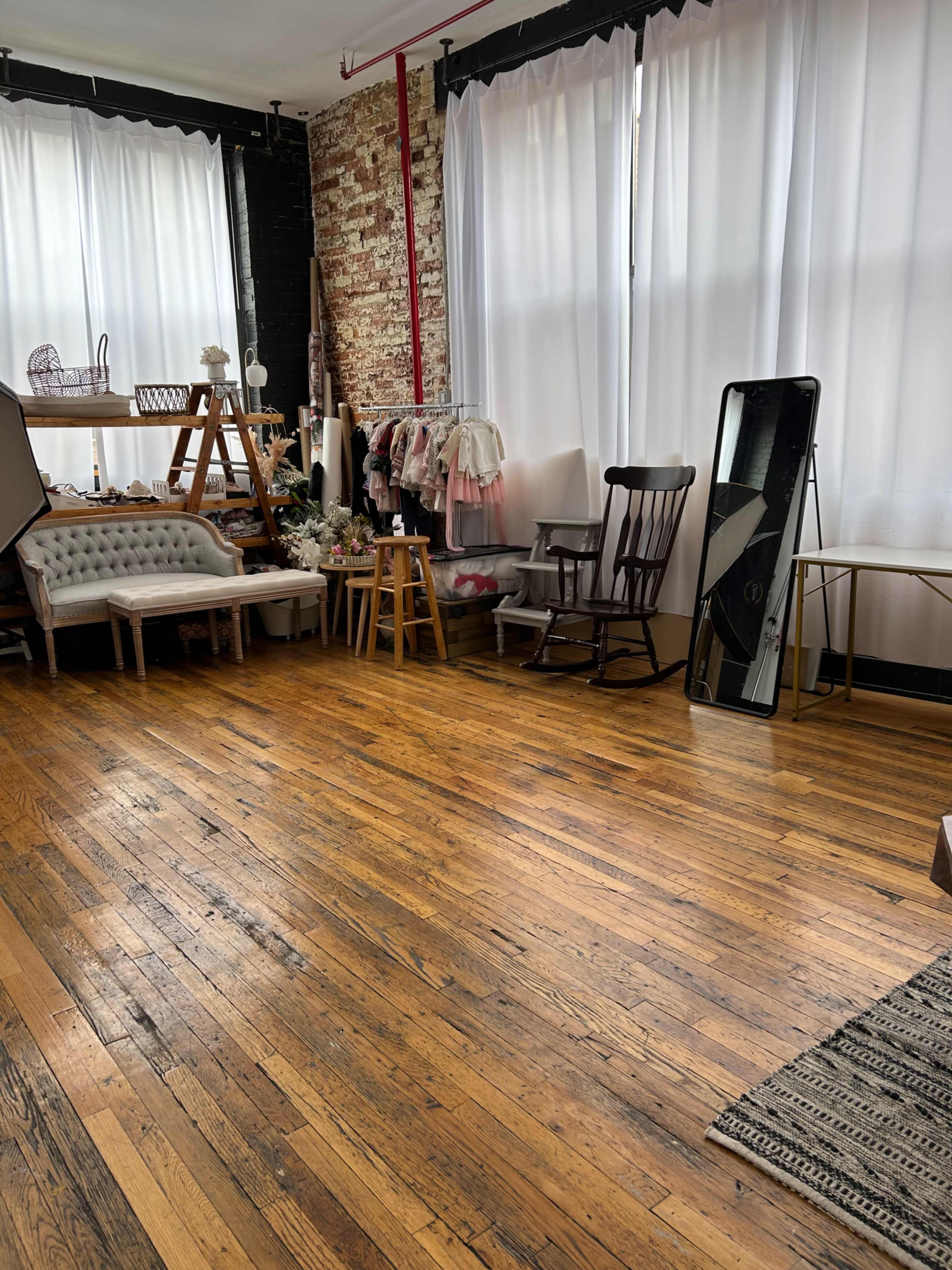 The room features hardwood floors, large windows with white curtains, and various furniture pieces including rocking chairs and a mirror.
