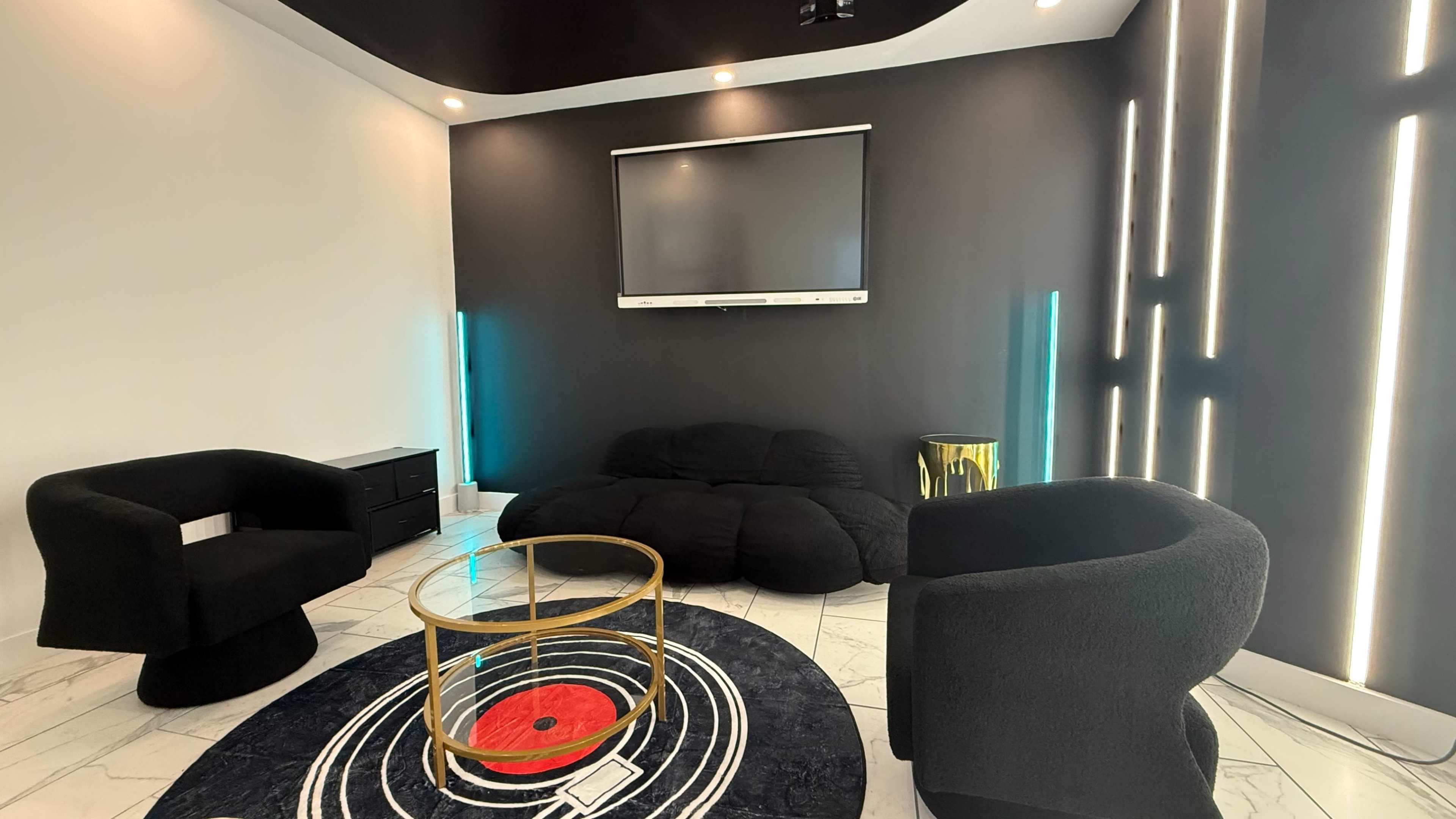 The image shows a modern lounge area with two black chairs, a round glass coffee table, a black couch, and a television mounted on a dark wall.