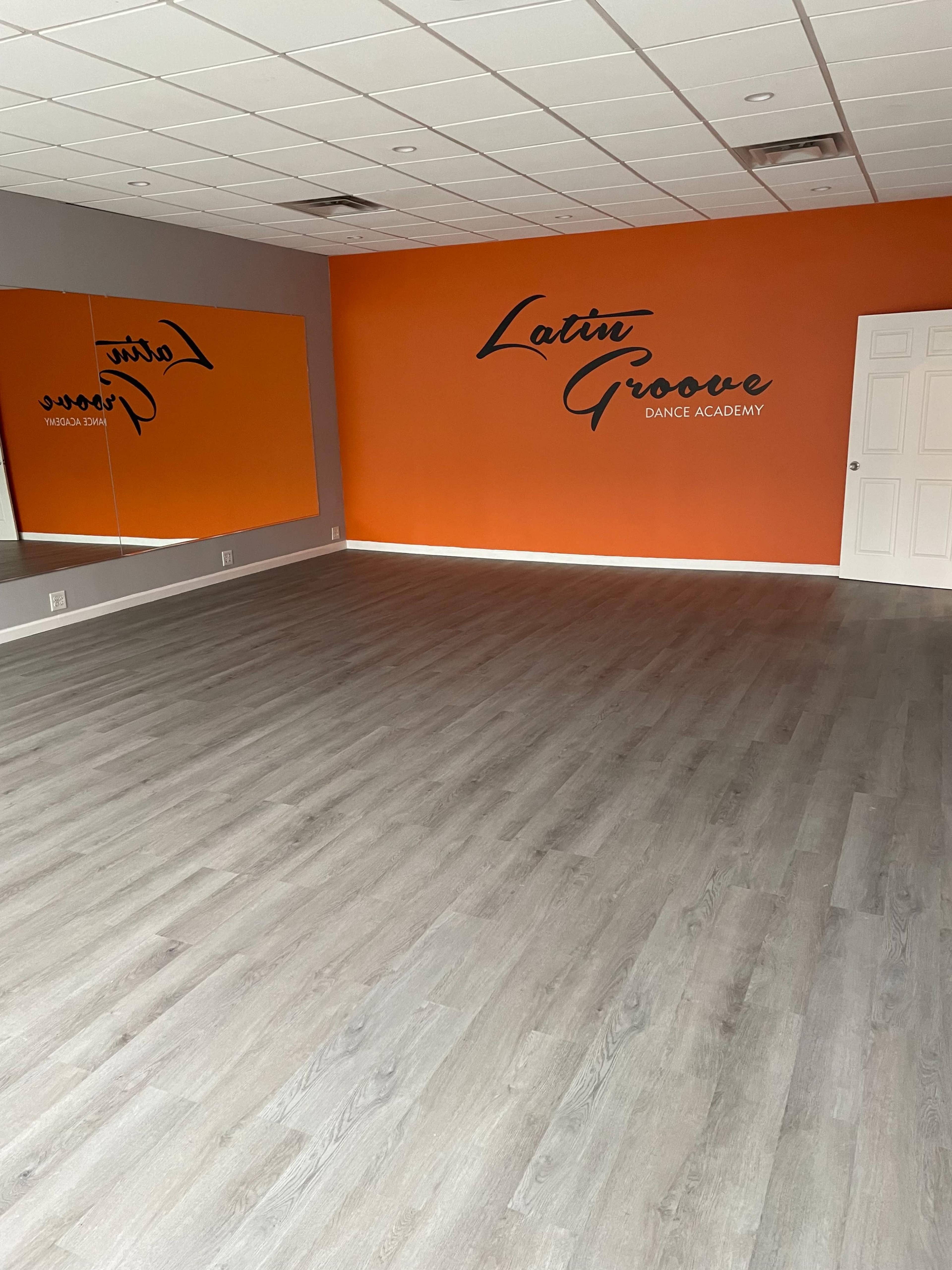 The image shows an empty dance studio with a gray floor, an orange accent wall featuring the text "Latin Groove Dance Academy," and a large mirror on one side.