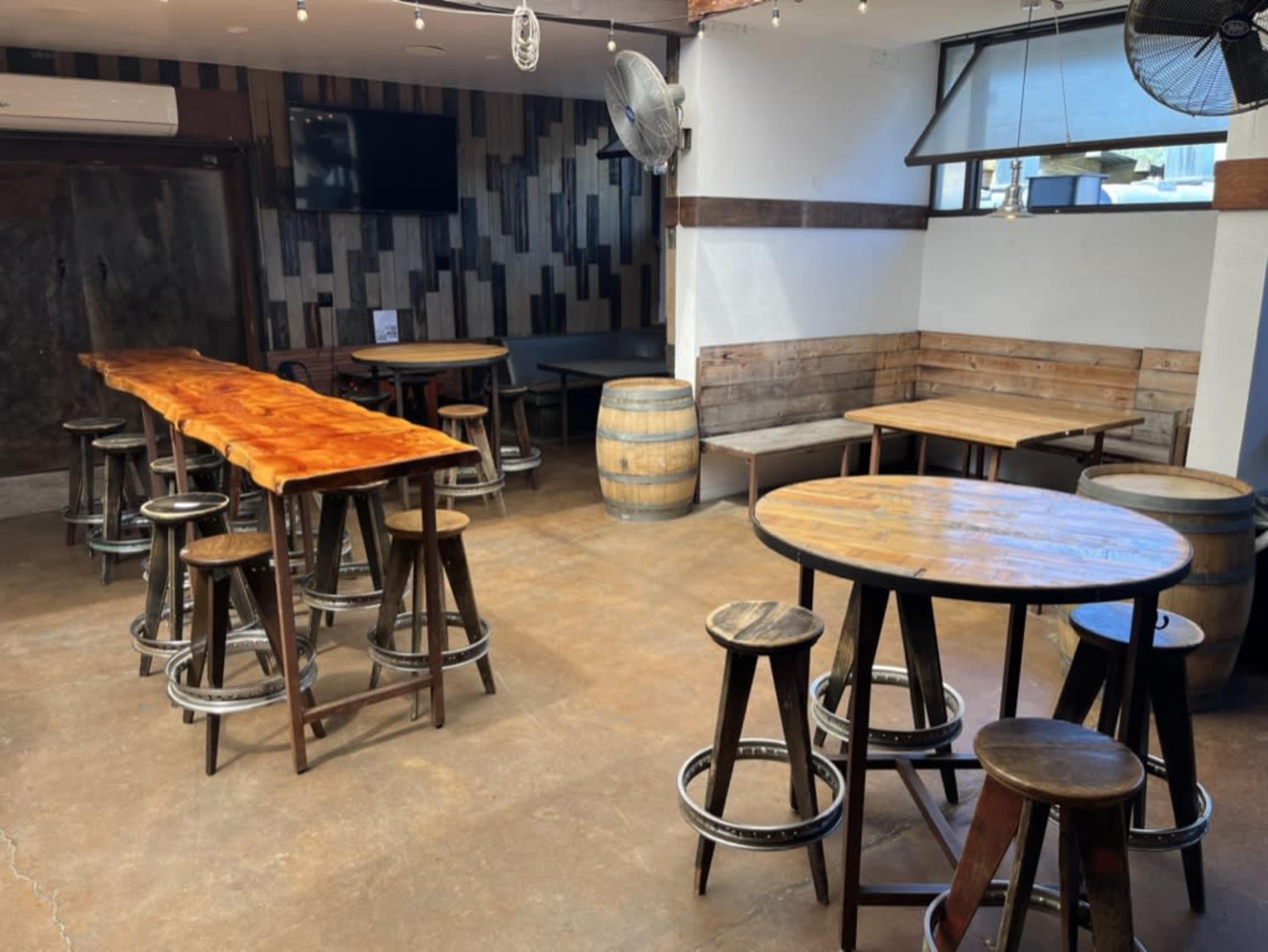 The image shows a spacious indoor area with wooden tables and stools, featuring a bar-style table with a shiny surface, surrounded by various seating options and decor elements.