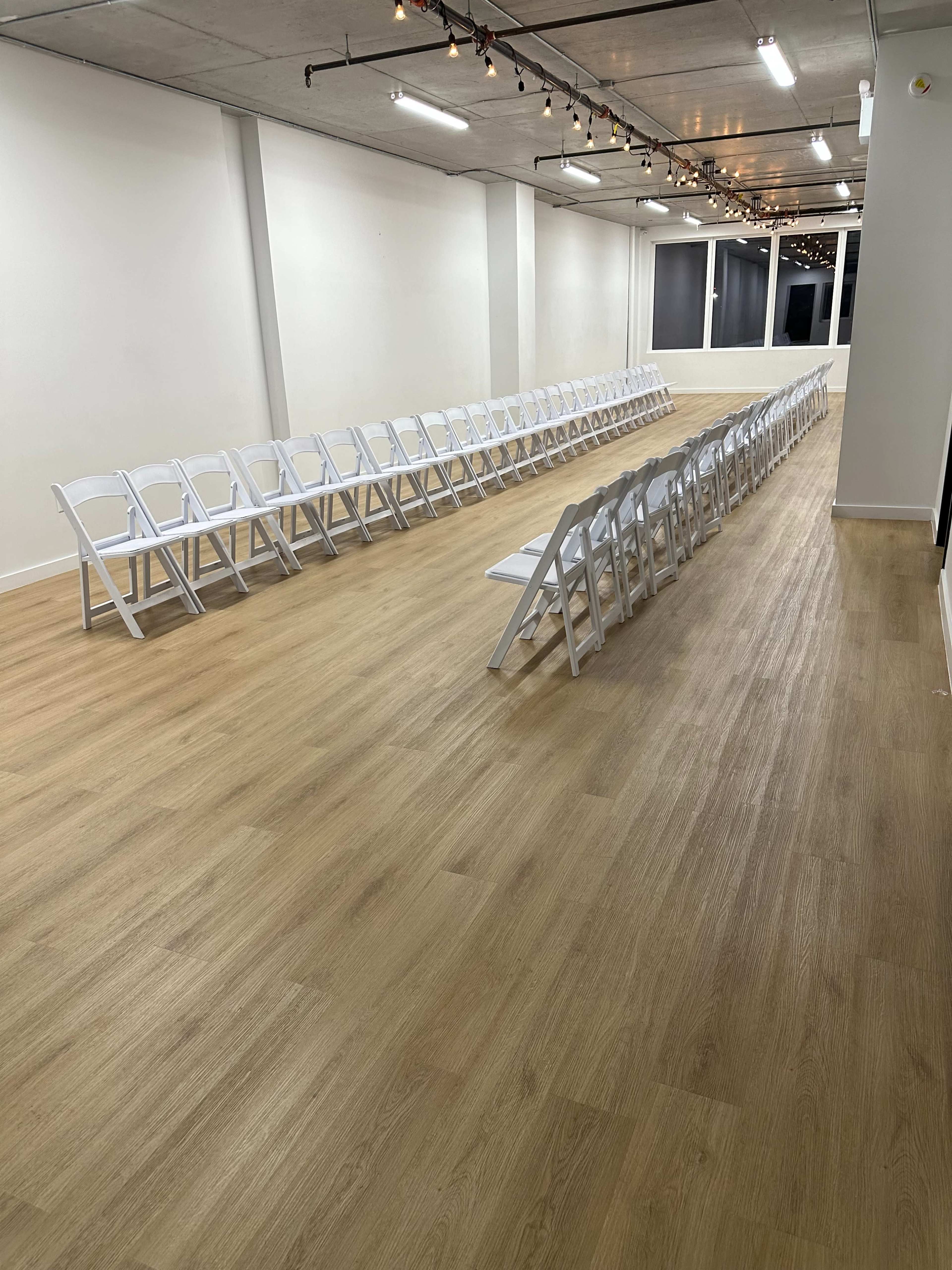 A well-lit room features two rows of white folding chairs arranged facing each other on a wooden floor.