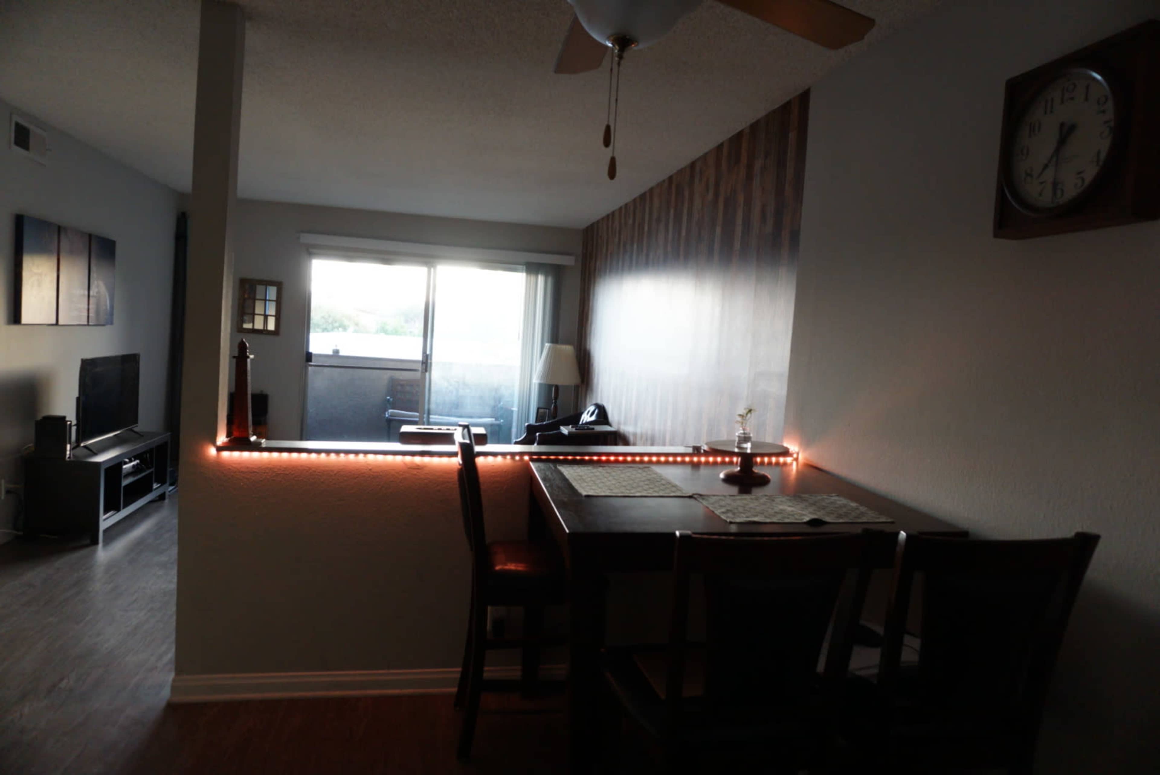 A dining area features a table with chairs next to a window overlooking a balcony, illuminated by LED lights along the wall.
