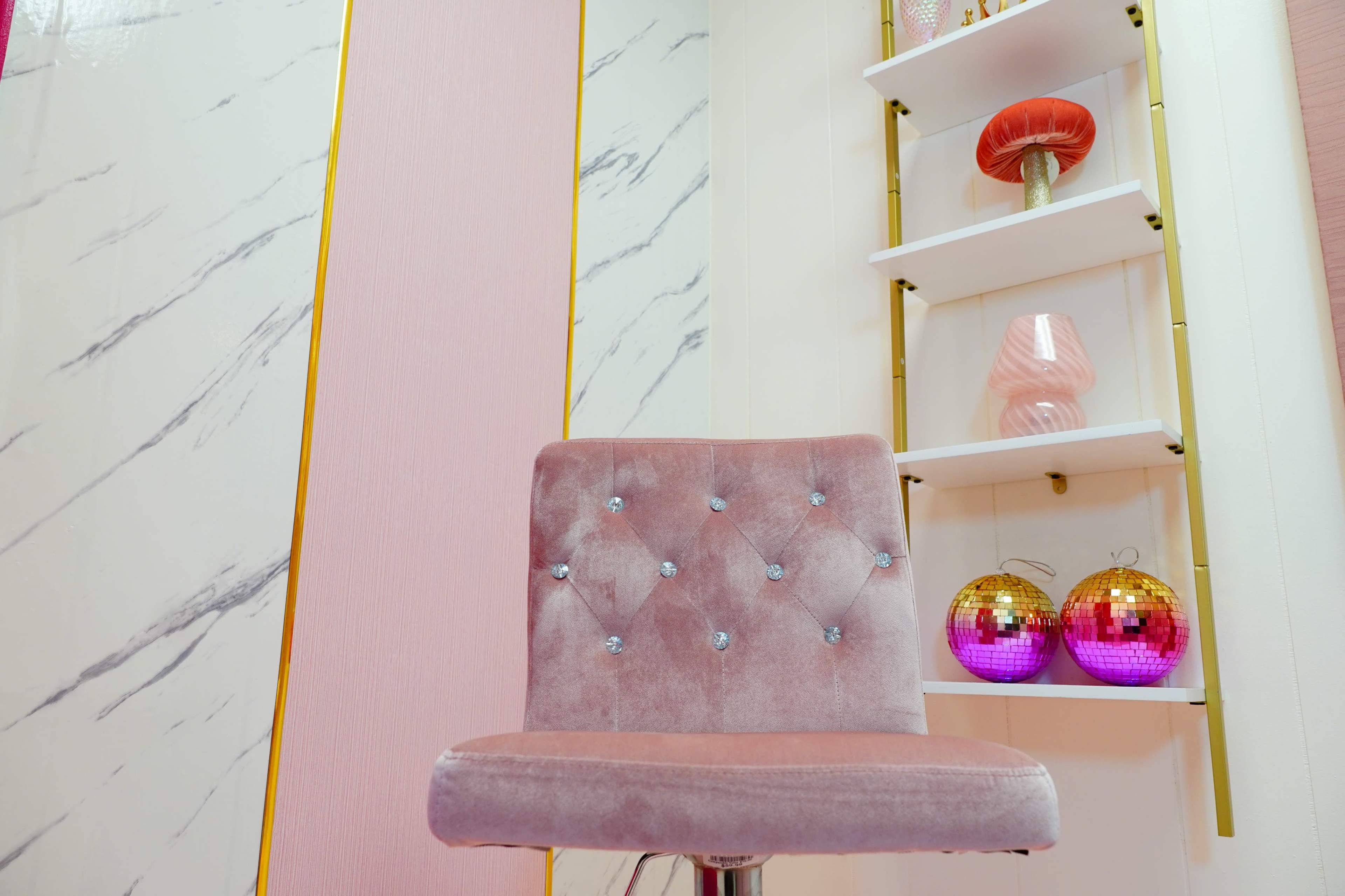 A pink cushioned chair with diamond tufting in front of a shelf displaying decorative items, including colorful ornaments and lamps.