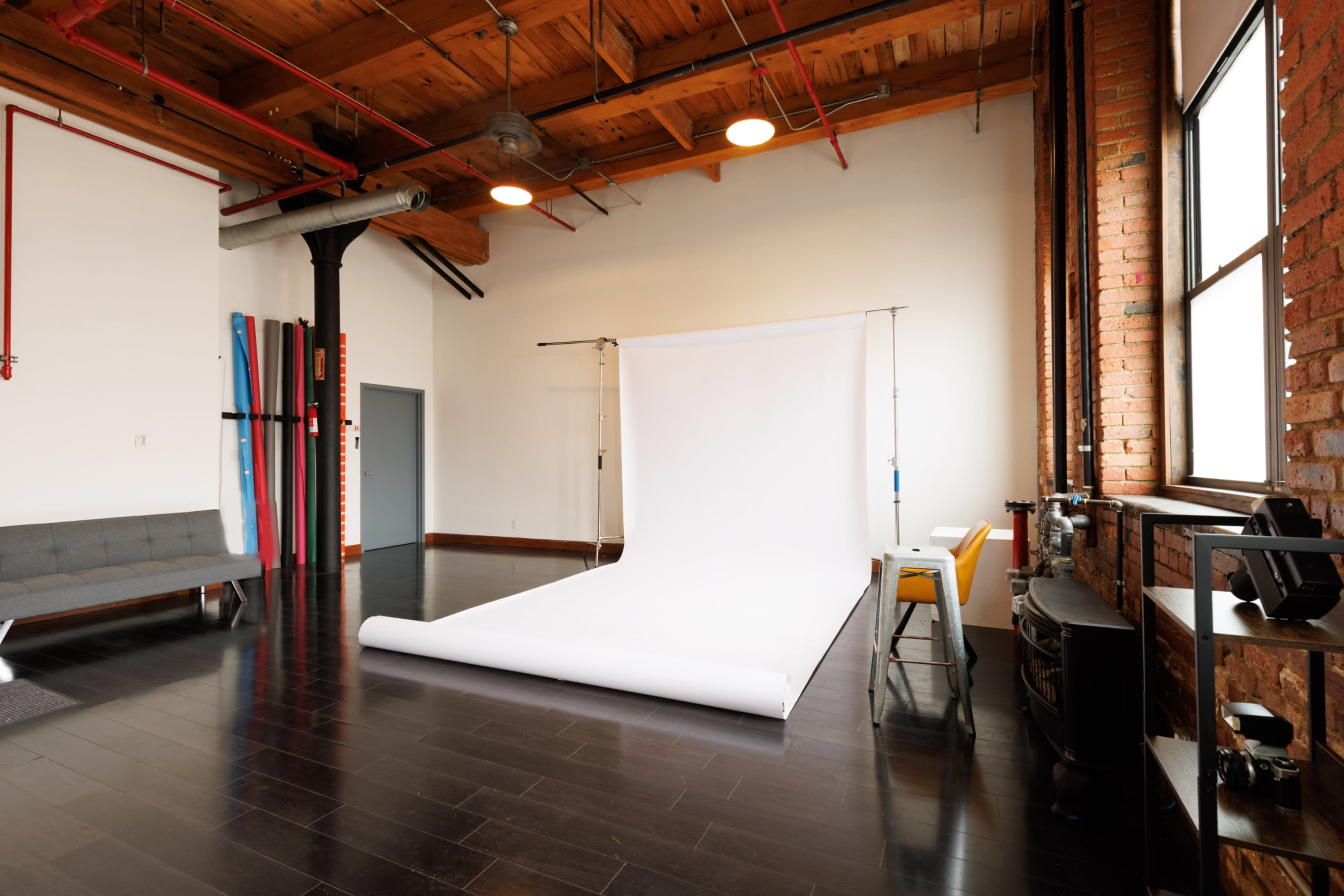 The image shows a spacious photography studio featuring a white backdrop, wooden ceilings, and large windows letting in natural light.