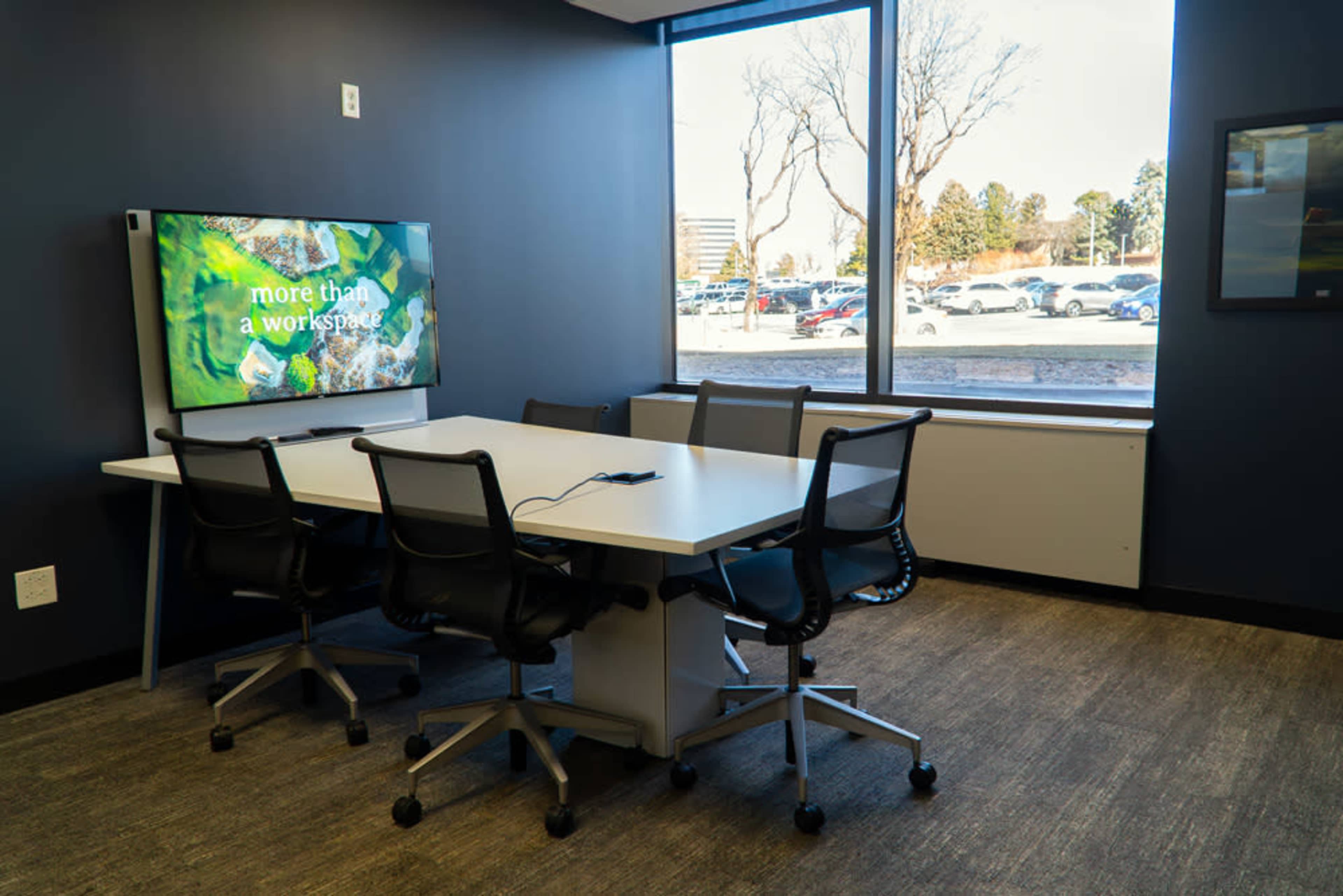 The image shows a small meeting room featuring a table with chairs, a television screen displaying text, and large windows with a view of a parking lot and trees outside.