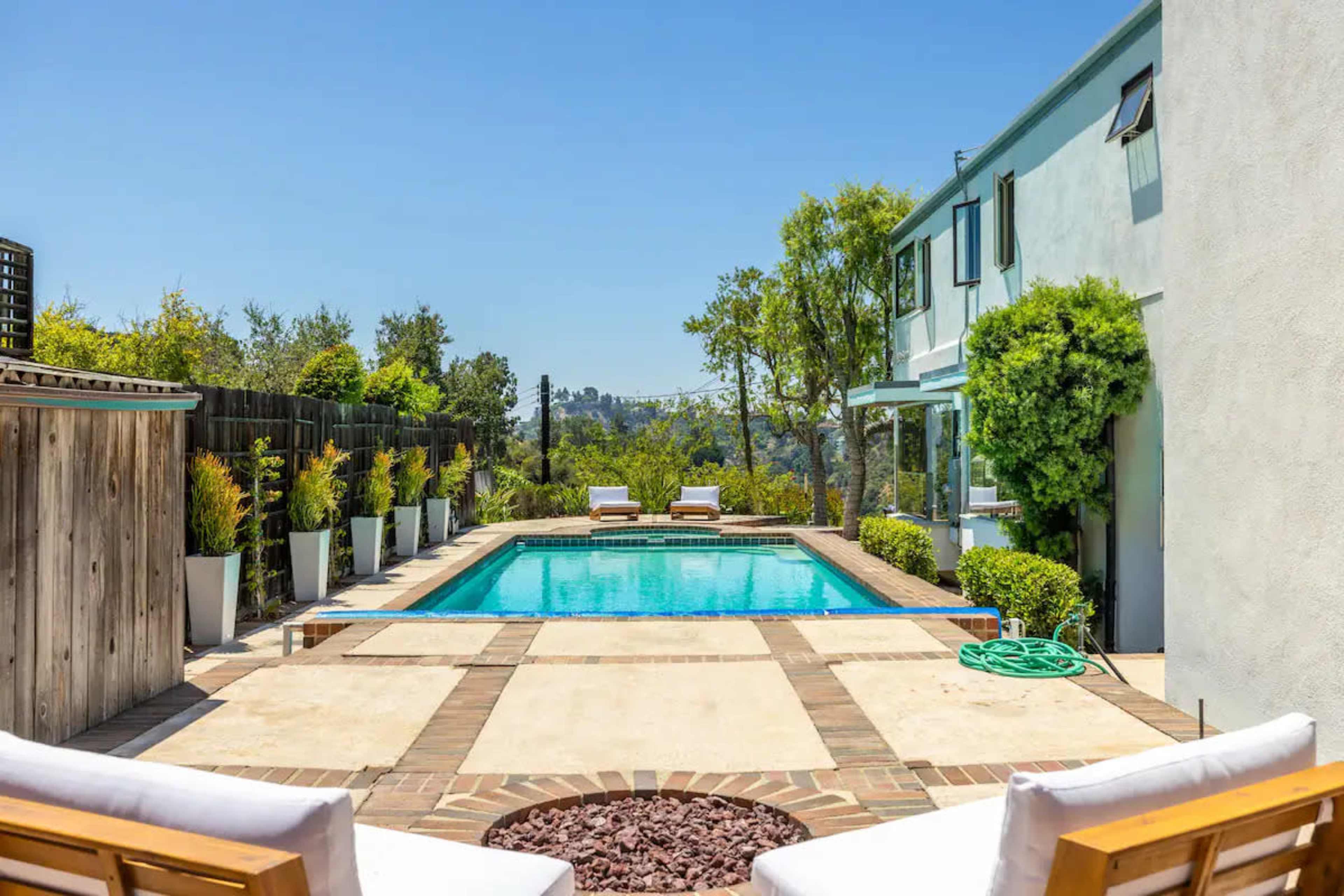 The scene shows a serene outdoor area featuring a rectangular swimming pool surrounded by lounge chairs and potted plants, with a house visible to the side.