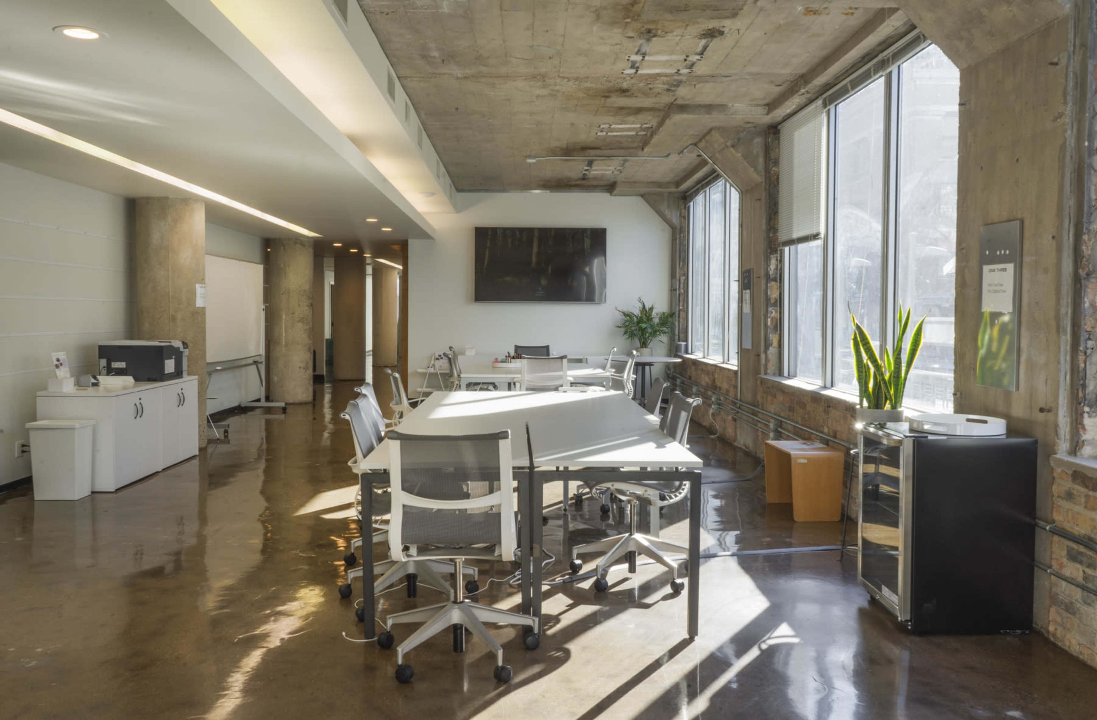 The image shows a modern, open office space with large windows, a long table surrounded by chairs, and various office furnishings along the walls.