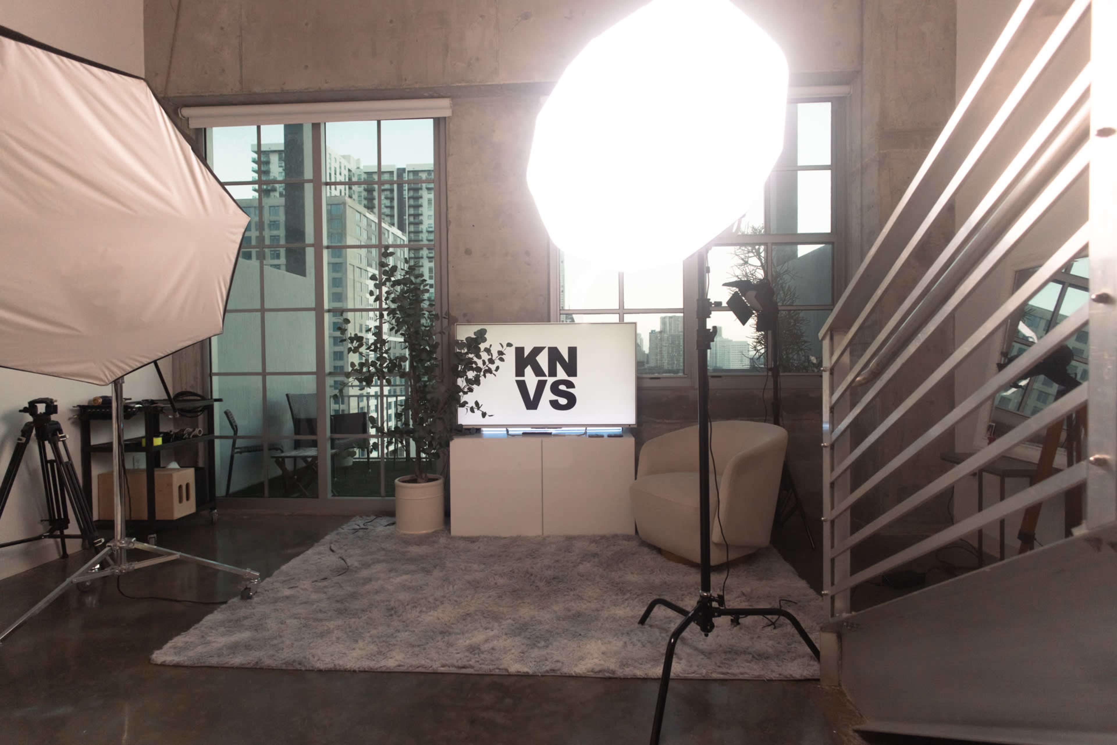The image shows a modern studio setup with a large light source, a softbox, and a display screen featuring the text "KN VS" against a backdrop of large windows.