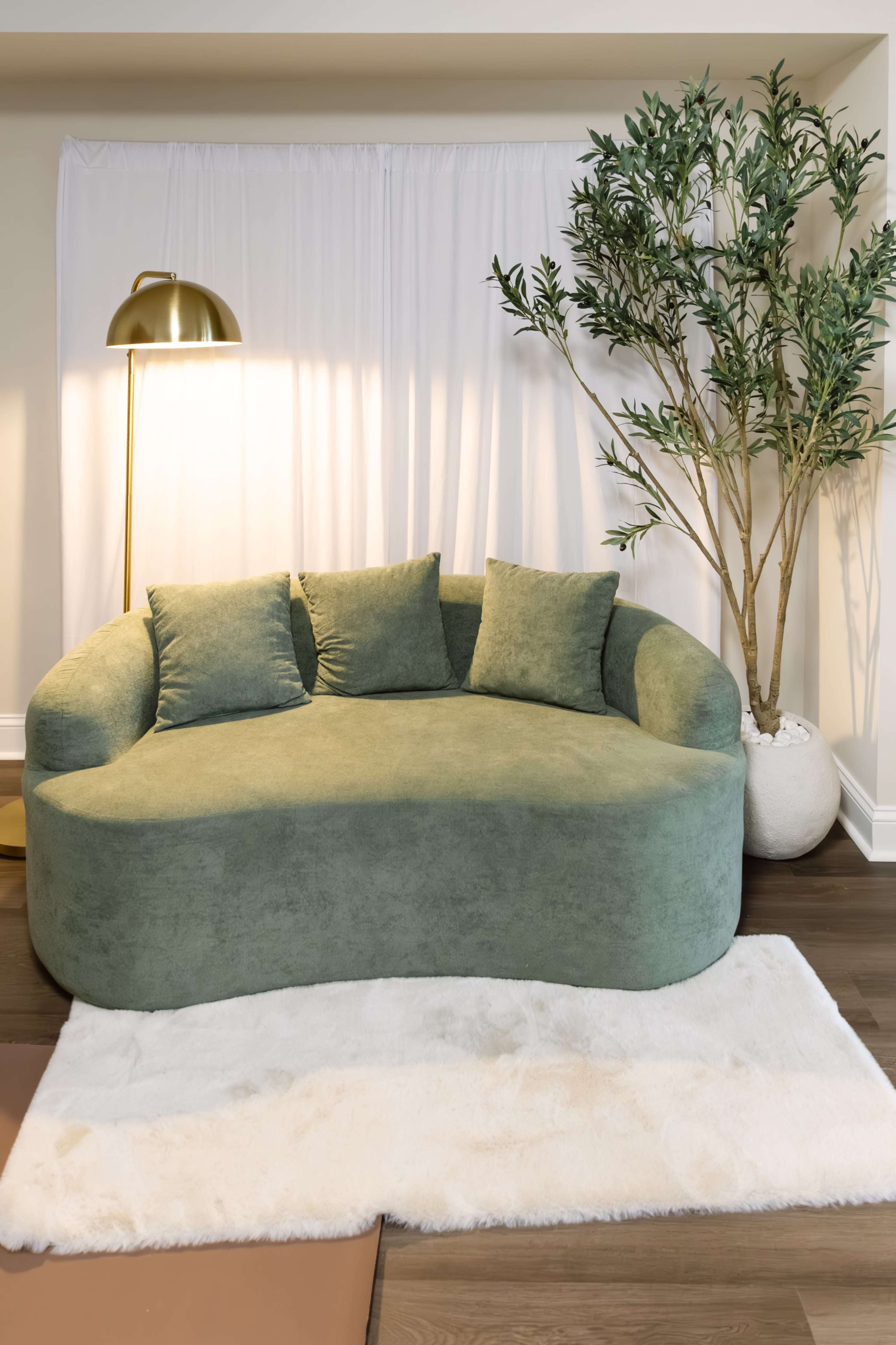 A curved green sofa with three cushions is positioned near a floor lamp and a potted plant in a softly lit room.