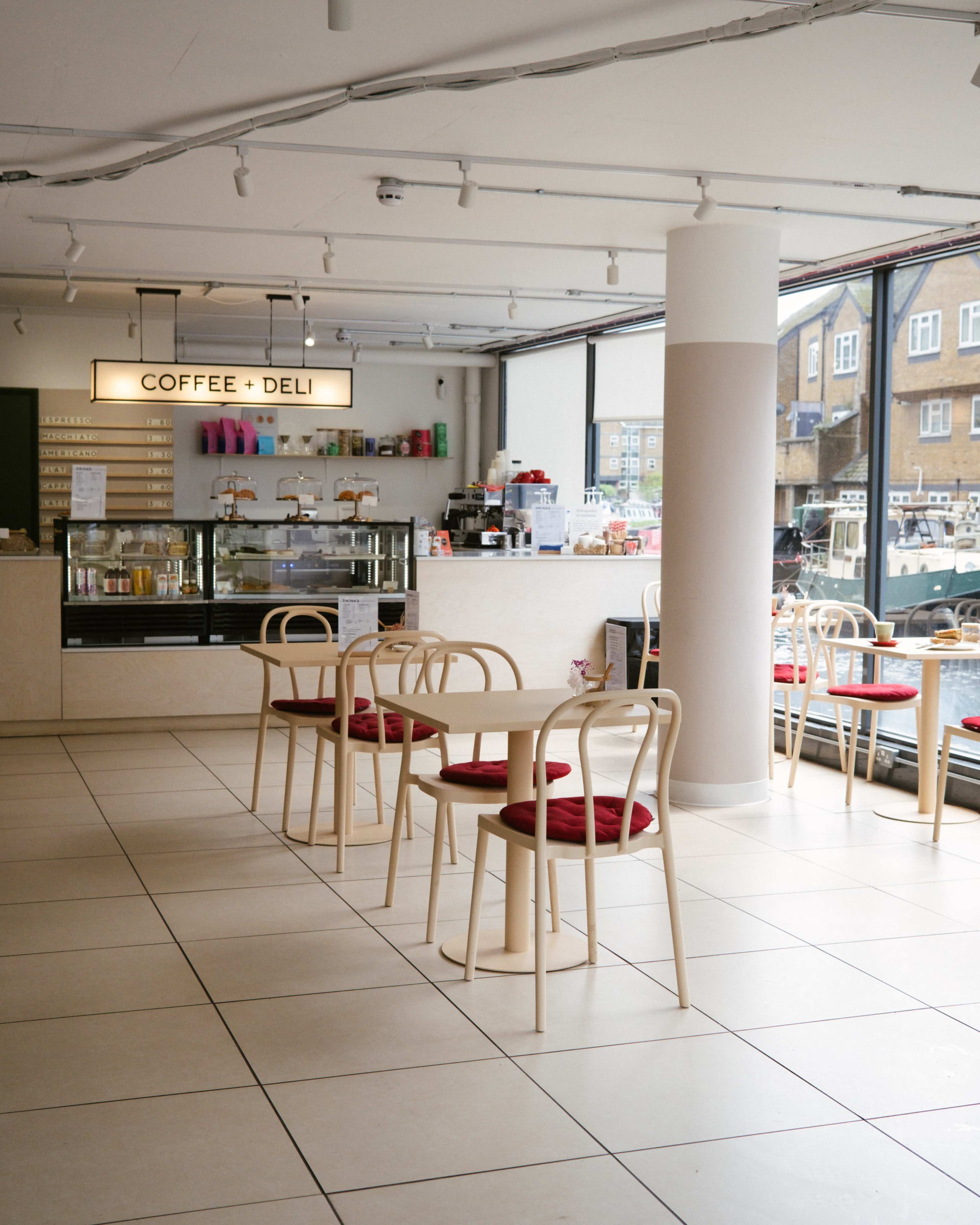 A modern coffee shop features light-colored furniture, a counter with beverages displayed, and large windows that overlook the street.