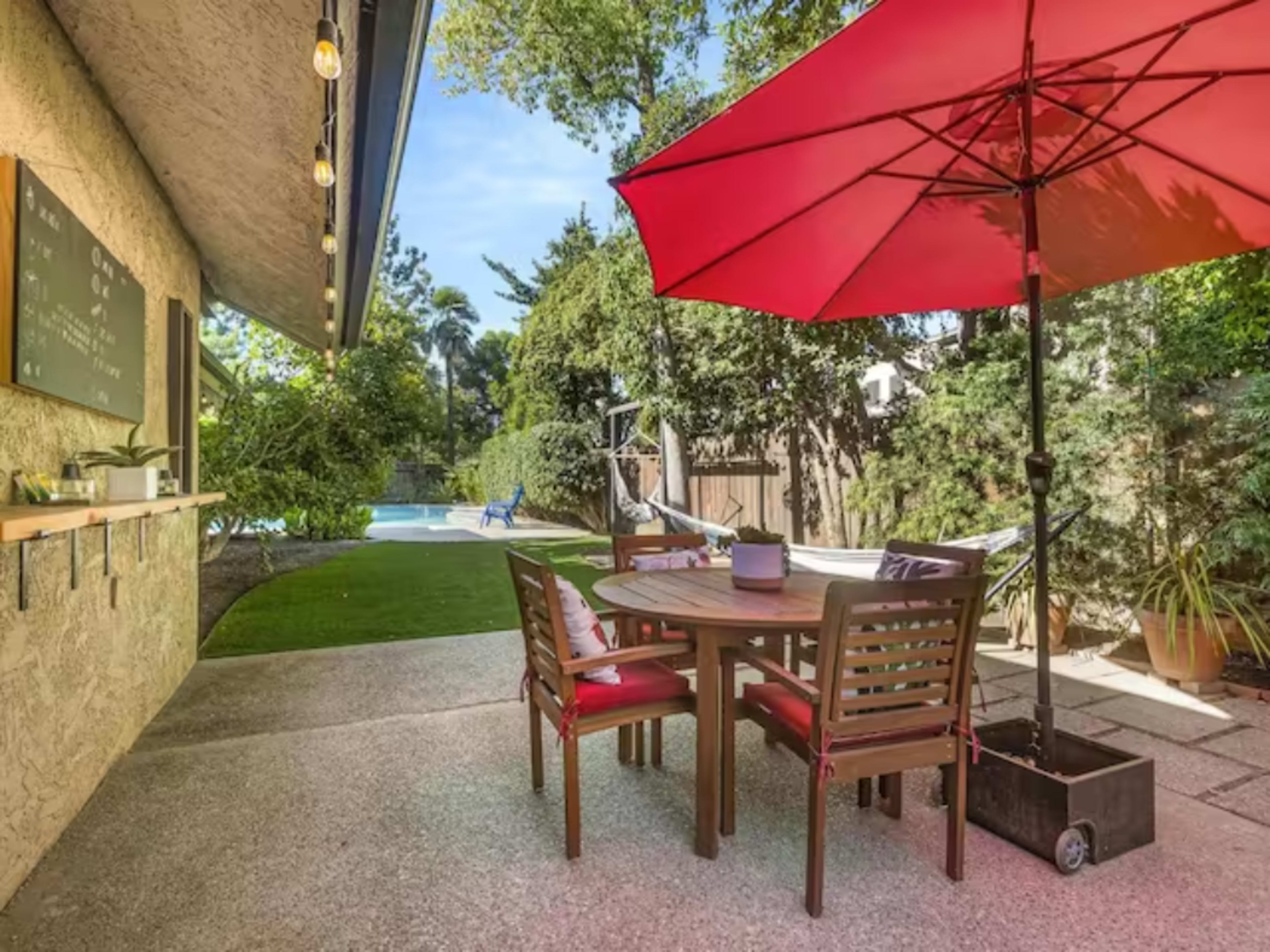A patio area features a round wooden dining table with chairs, shaded by a red umbrella, overlooking a grassy yard and swimming pool.