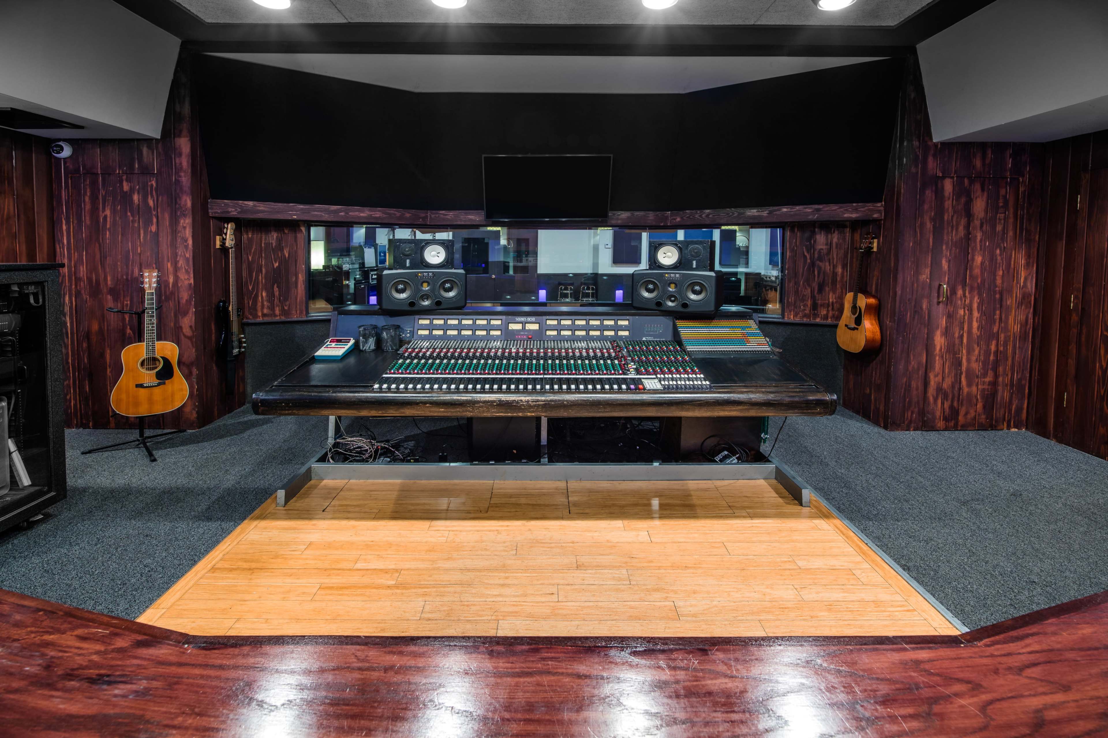 A music recording studio featuring a large mixing console, monitor speakers, an acoustic guitar on the wall, and wooden paneling.