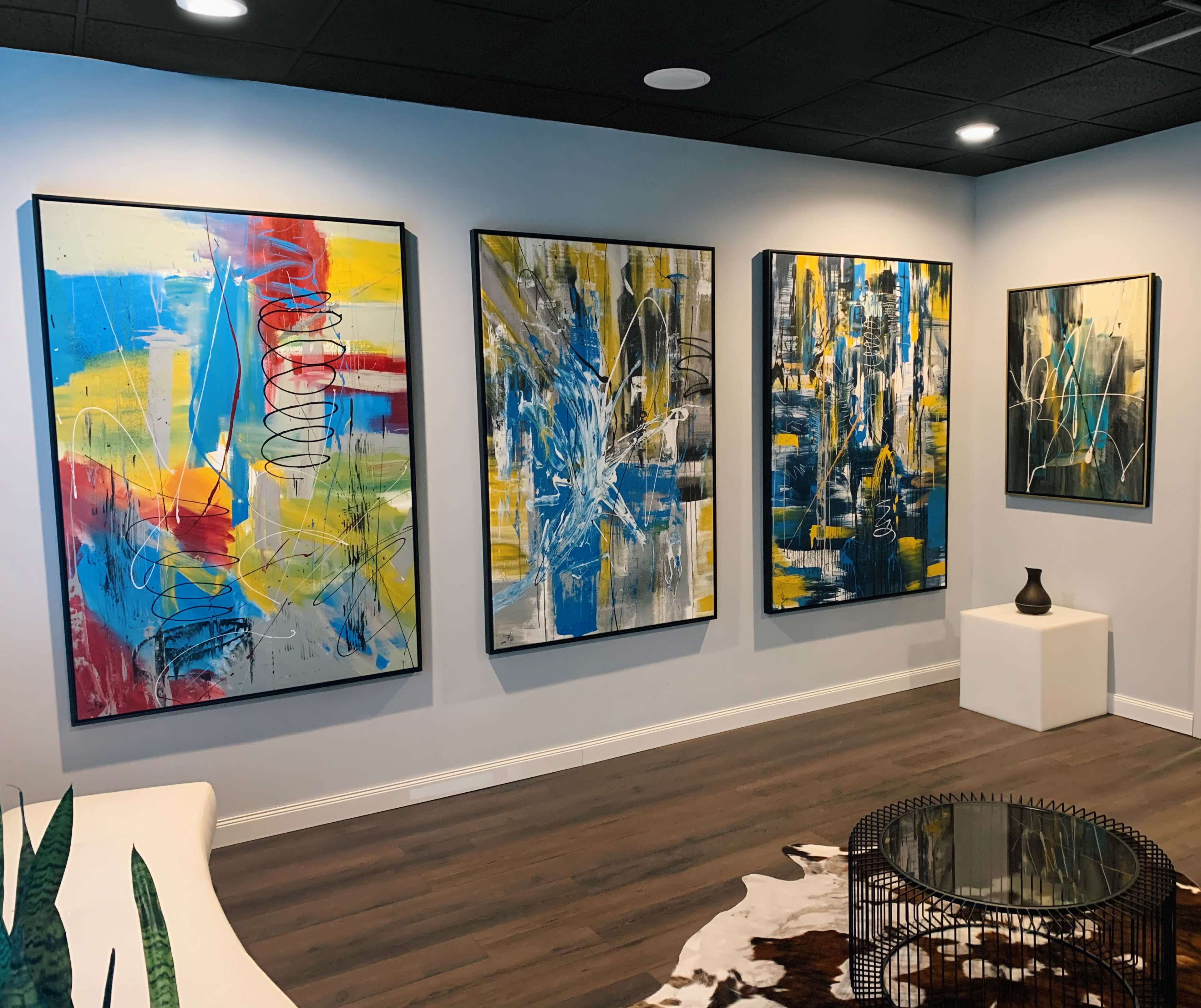 A modern art gallery featuring four large abstract paintings on a light-colored wall, with a glass coffee table and a decorative object on a pedestal in the center of the room.