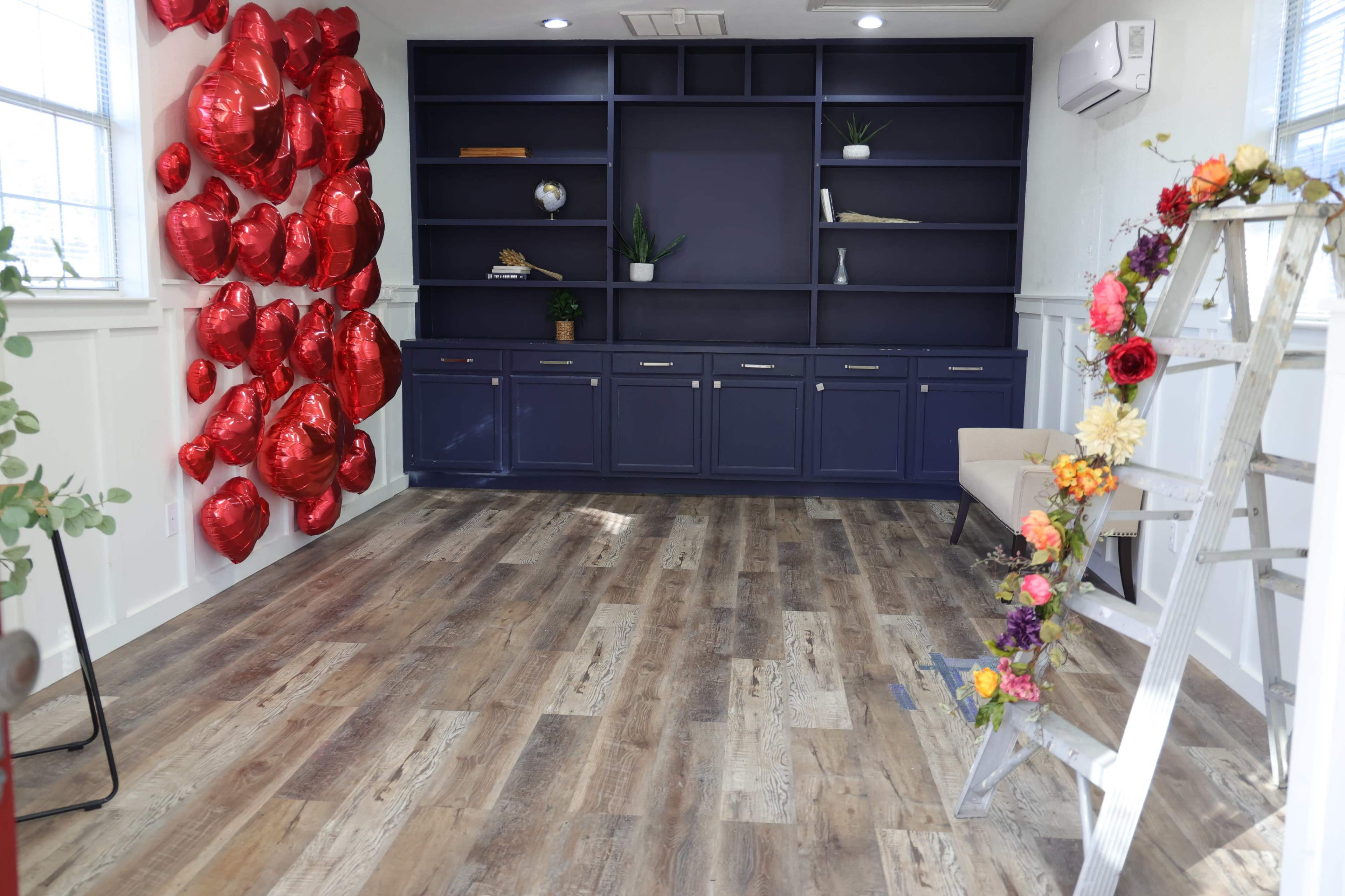 The image shows a room with a navy blue built-in shelving unit, red heart-shaped balloons on one wall, a wooden ladder adorned with flowers, and a beige chair.