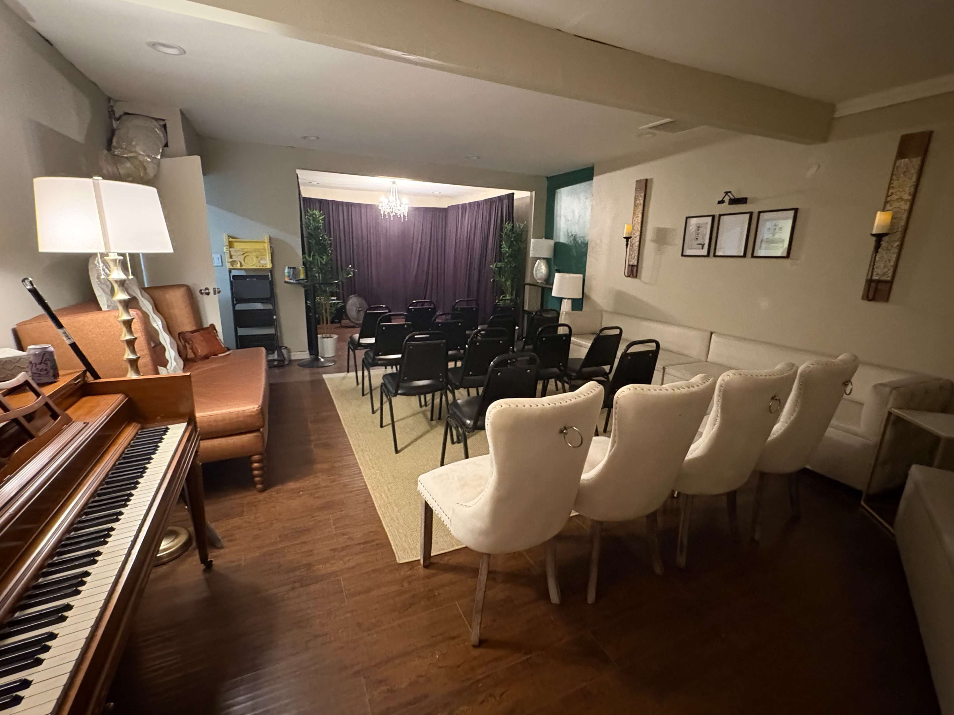 The image depicts a small room set up for a presentation or event, featuring a row of black chairs facing a stage area with a curtain and decorative plants, while a wooden piano is positioned on the left side.