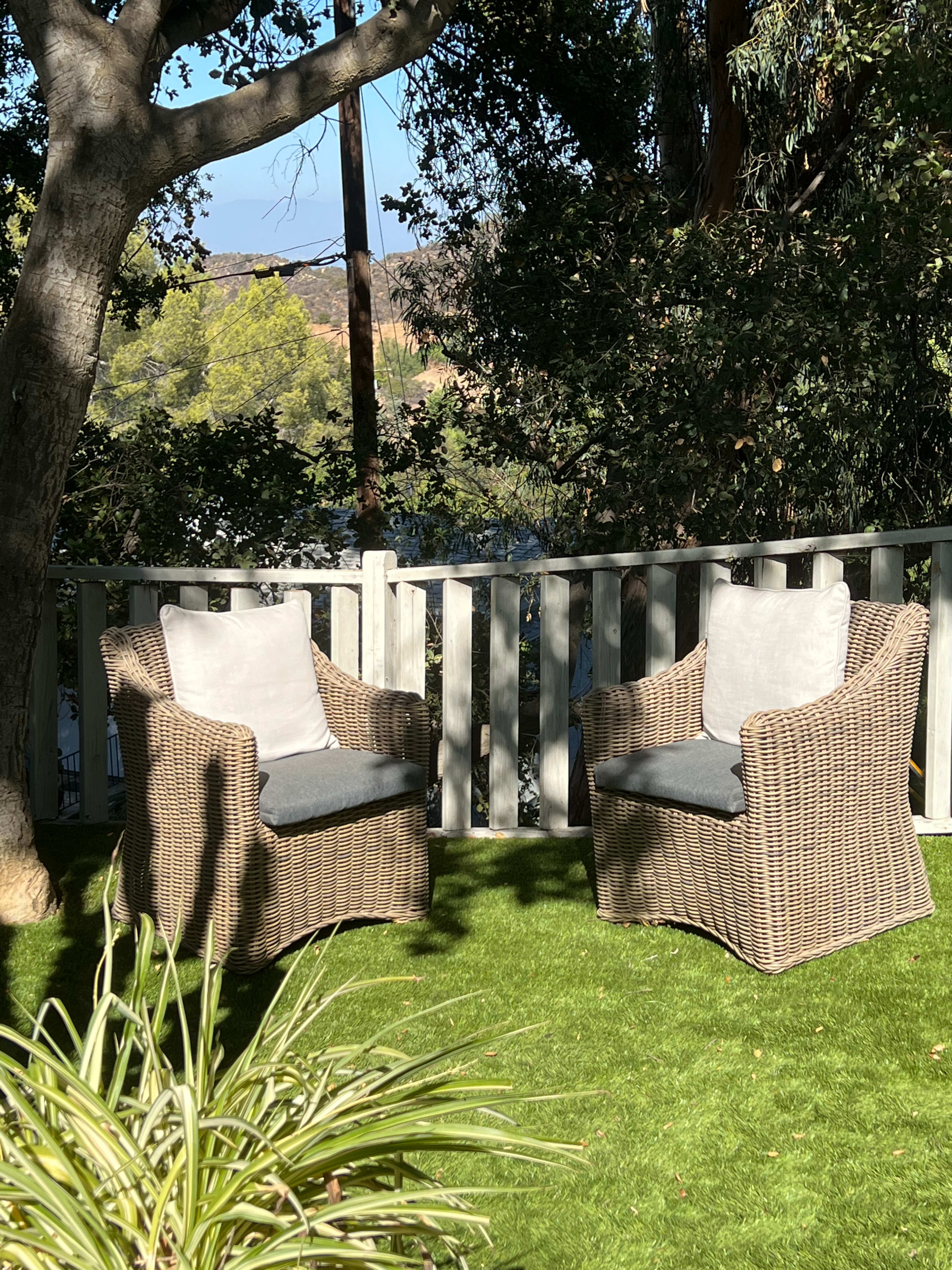 Two wicker chairs with cushions are positioned on a green lawn surrounded by a wooden fence and trees.