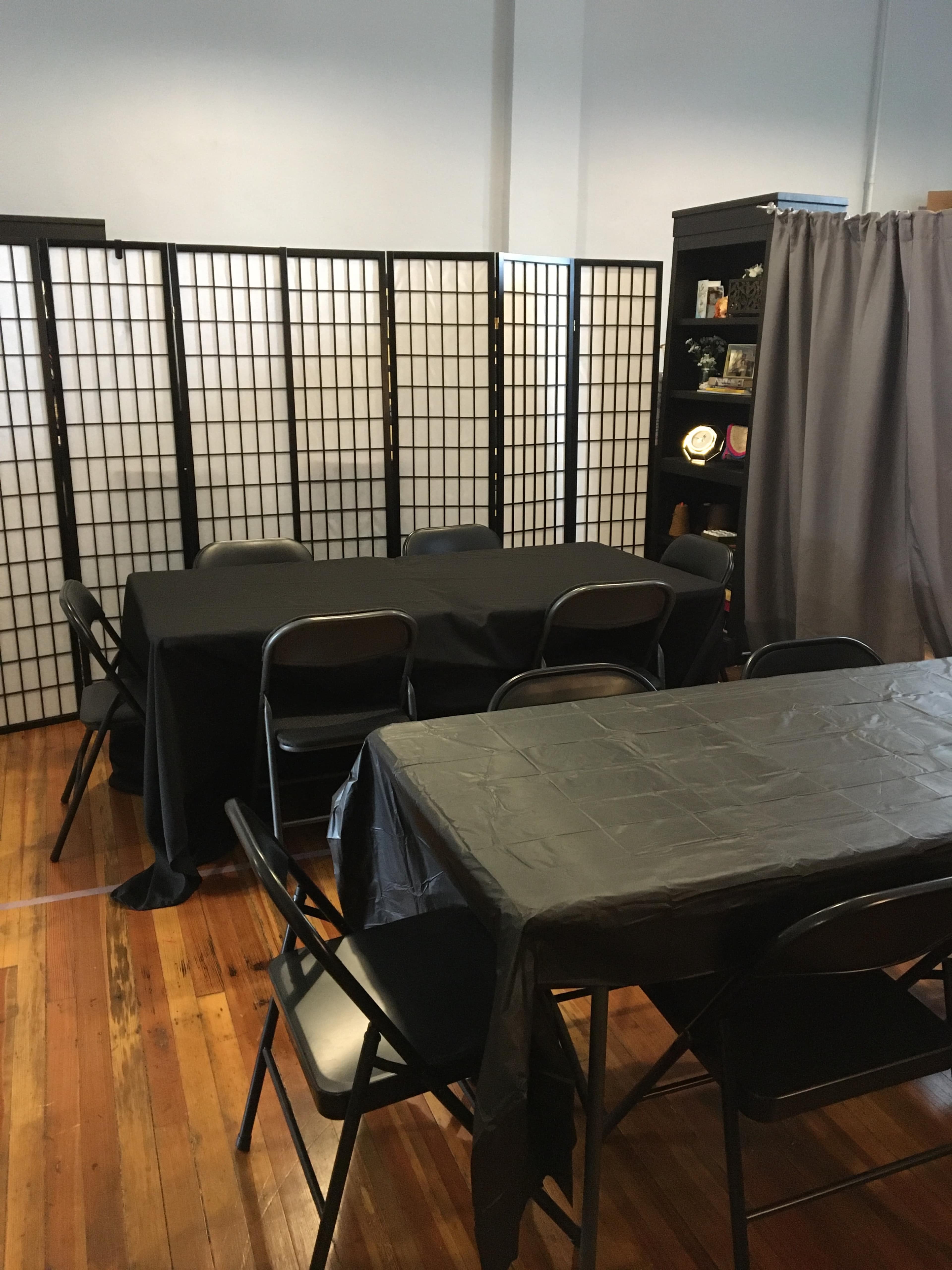 The room features two tables covered with black tablecloths surrounded by stacked black chairs, with folding screens and a curtain in the background.
