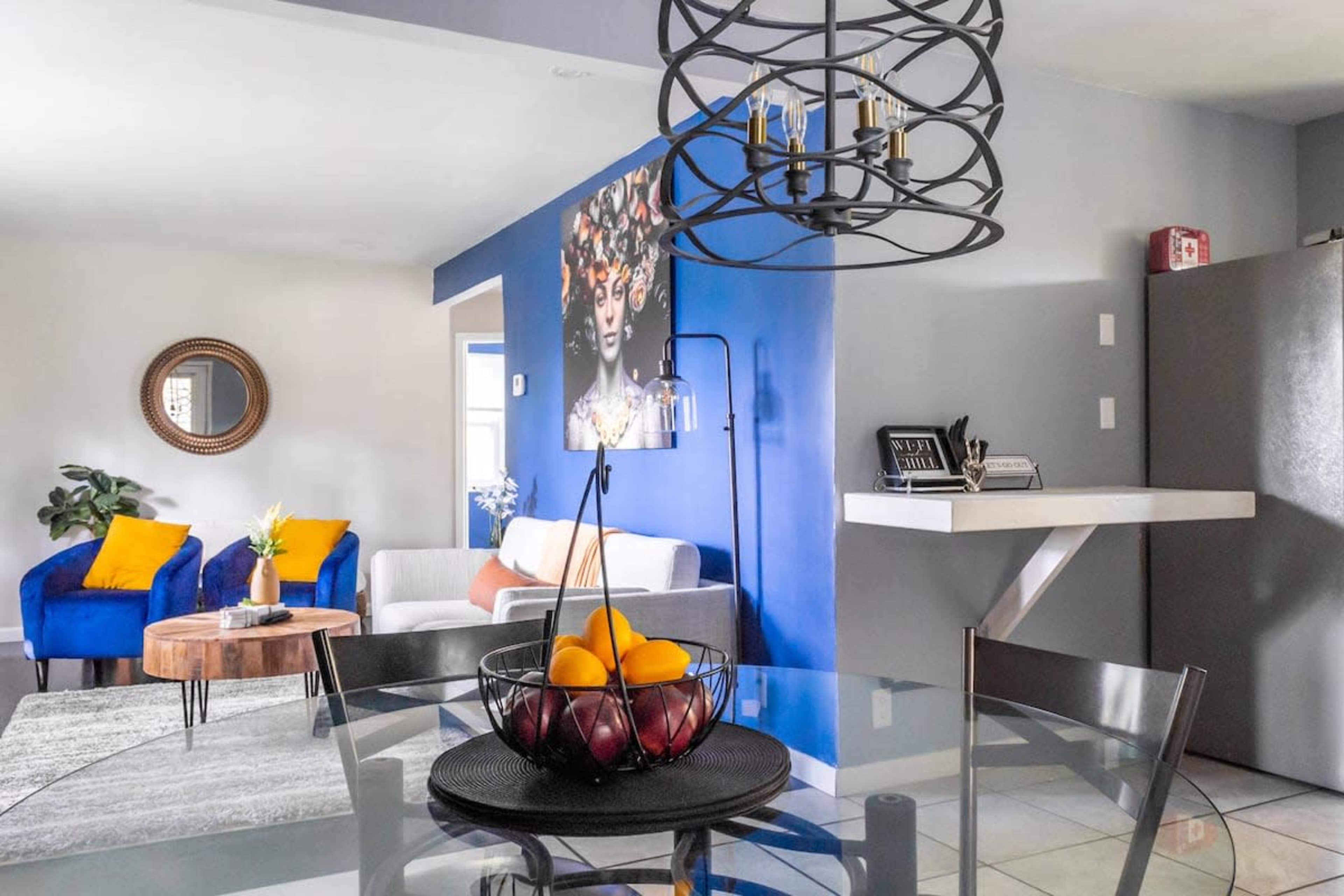 A modern living space features a round dining table with a fruit bowl, a gray sofa, blue accent chairs, and vibrant wall art.