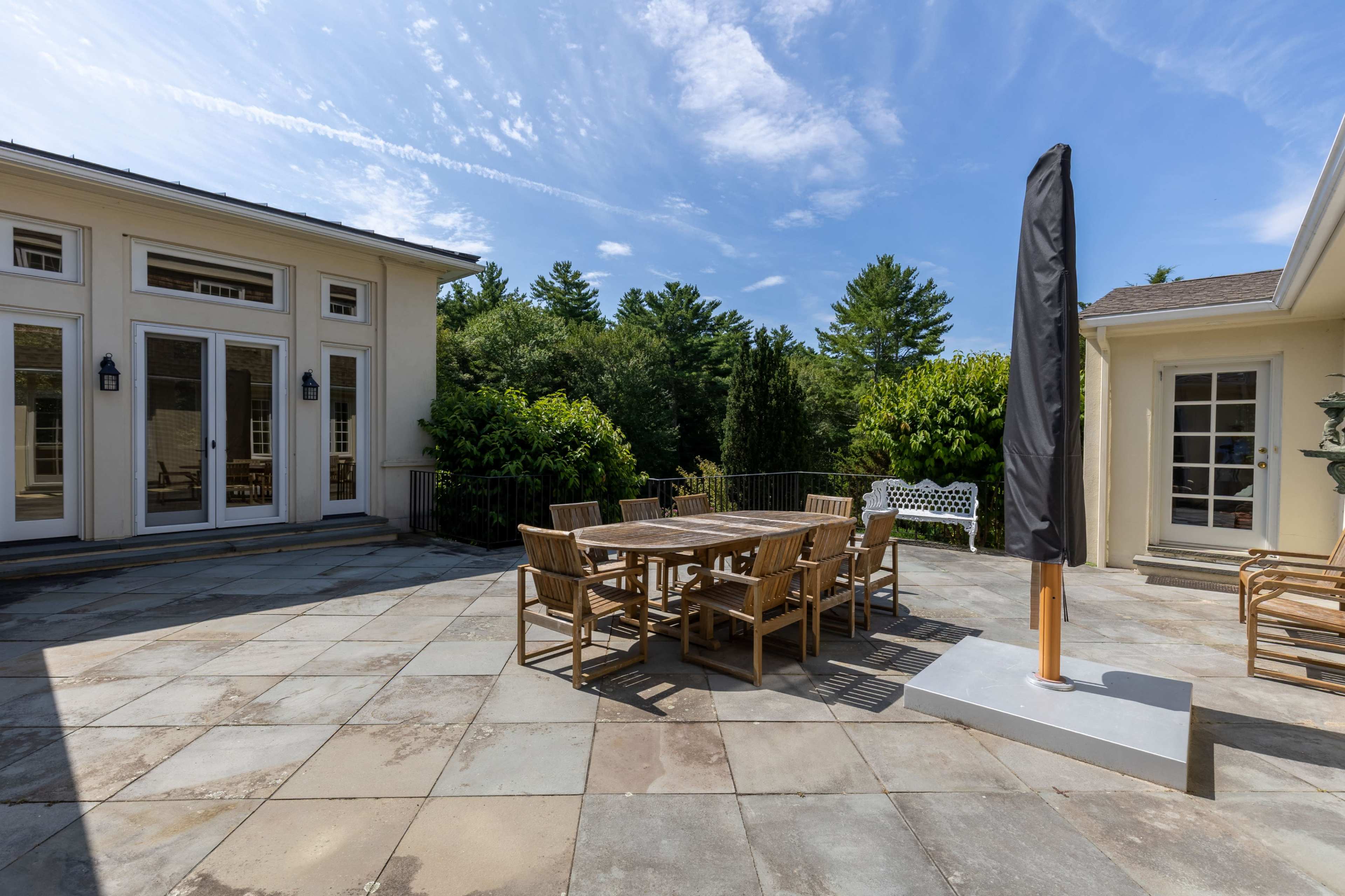 A sunlit patio features a wooden dining table surrounded by chairs, with a large umbrella and stone flooring, adjacent to a house and greenery.