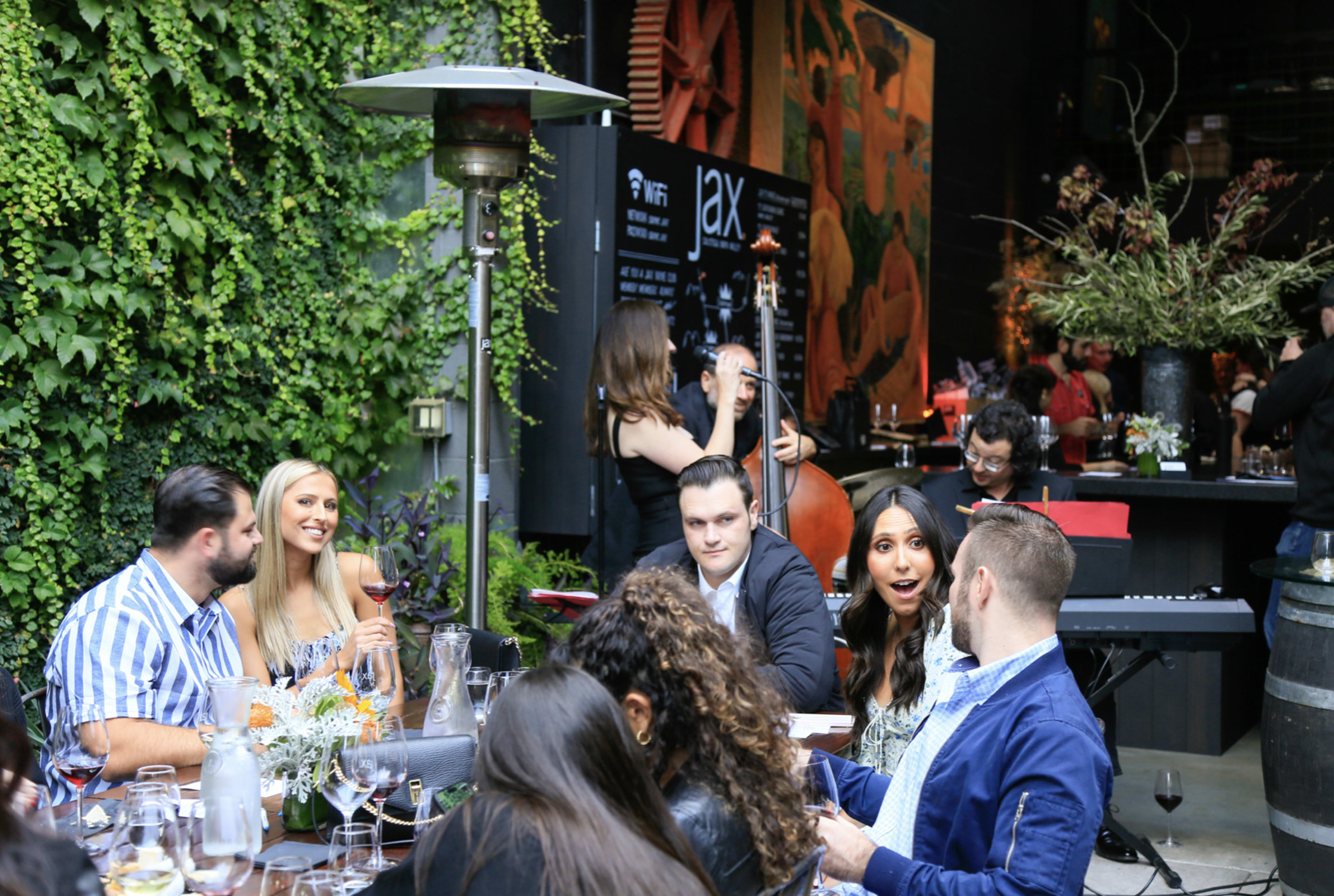 A group of people enjoys drinks and conversation in a lush, green outdoor dining area, with a musician playing in the background.