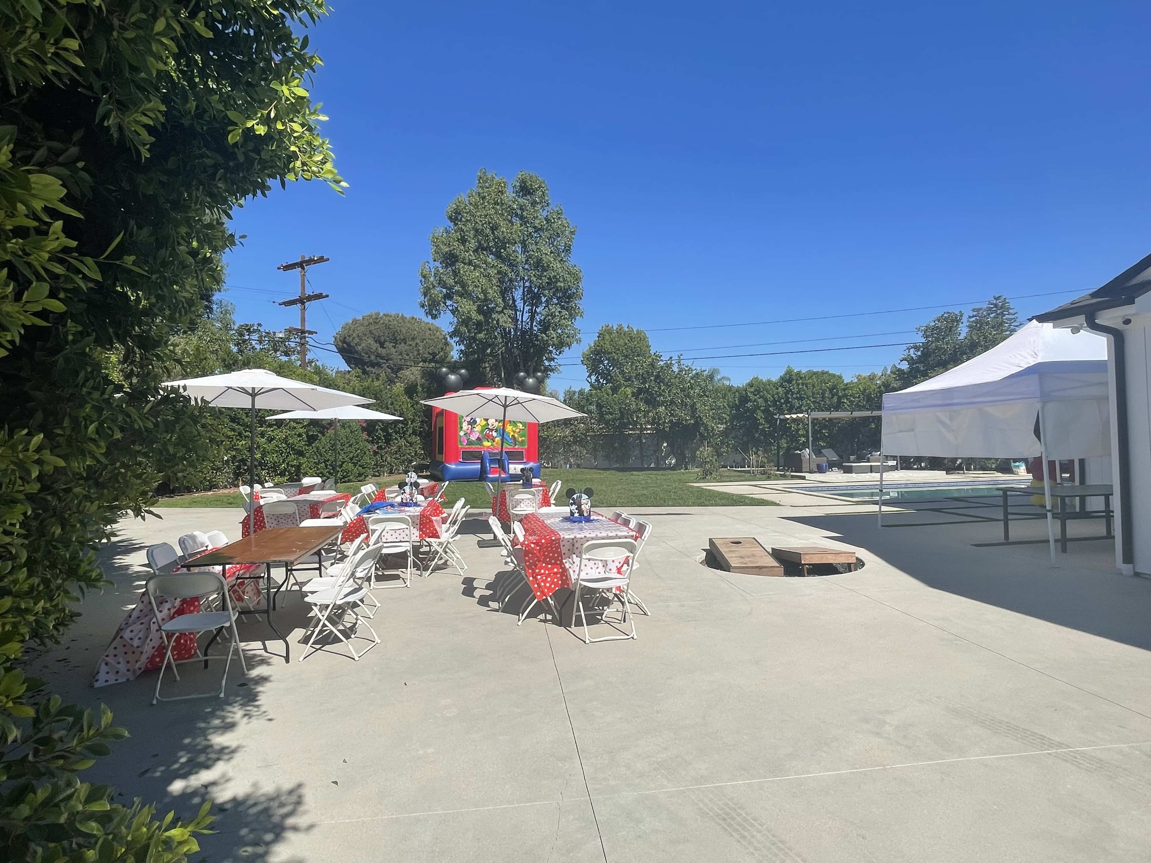 An outdoor party setting with tables covered in red and white tablecloths, shaded by umbrellas, and a bounce house in the background.