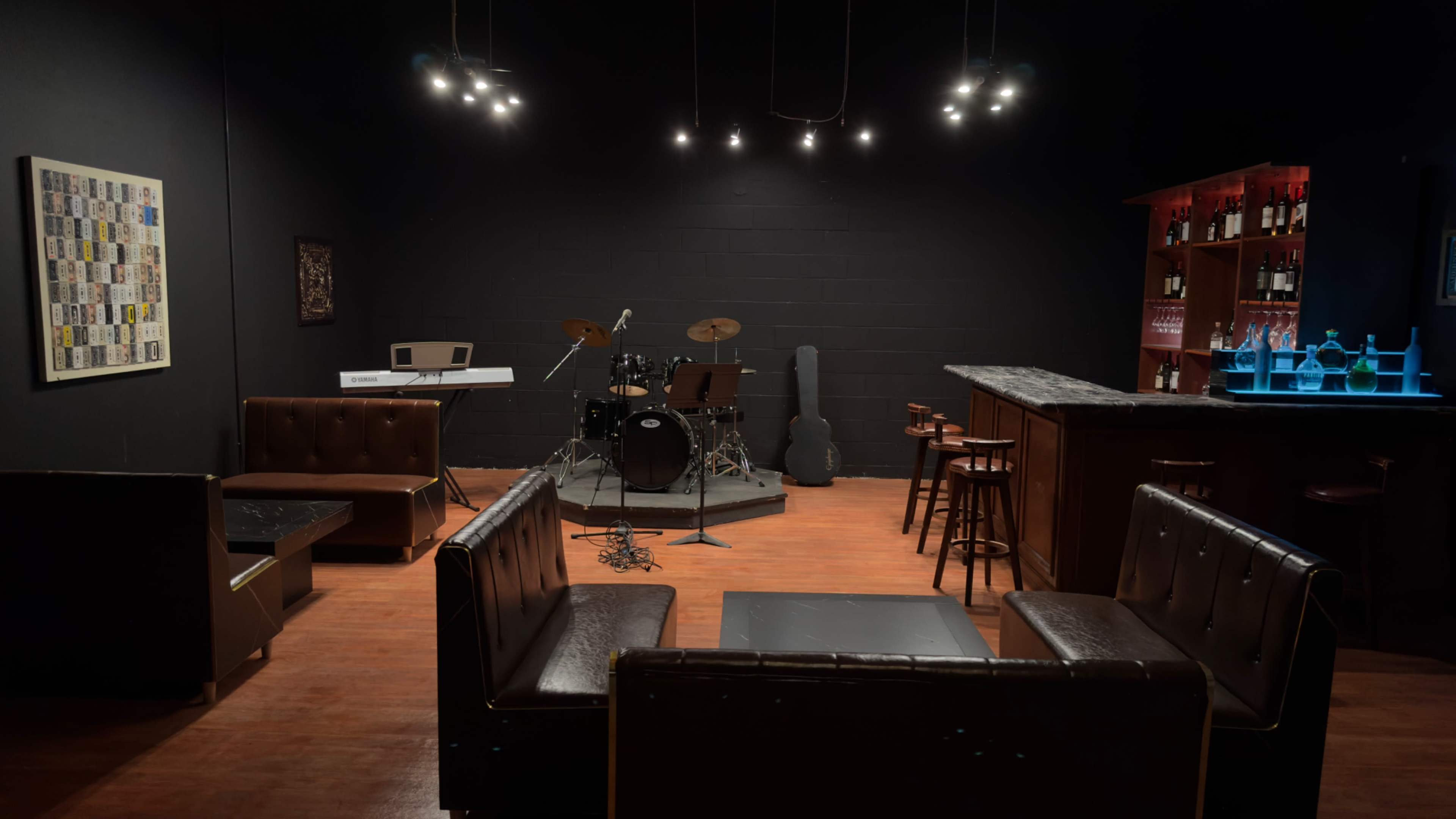 A dimly lit lounge area featuring a small stage with a drum set, a bar with bottles, and several leather couches arranged around a coffee table.