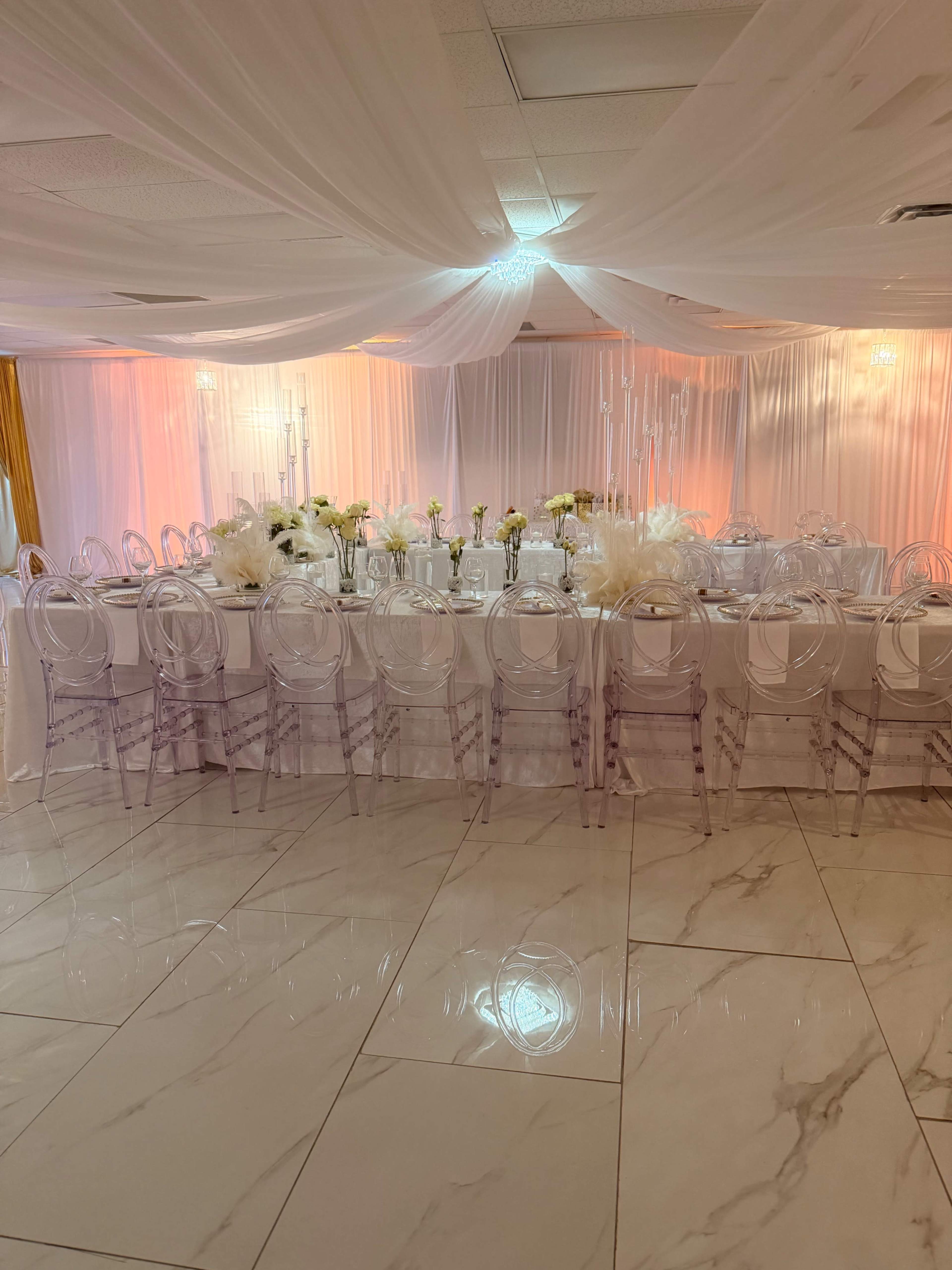 The image shows a banquet hall with elegantly arranged tables covered in white cloths, surrounded by transparent chairs, and adorned with flowers and decorative draping overhead.