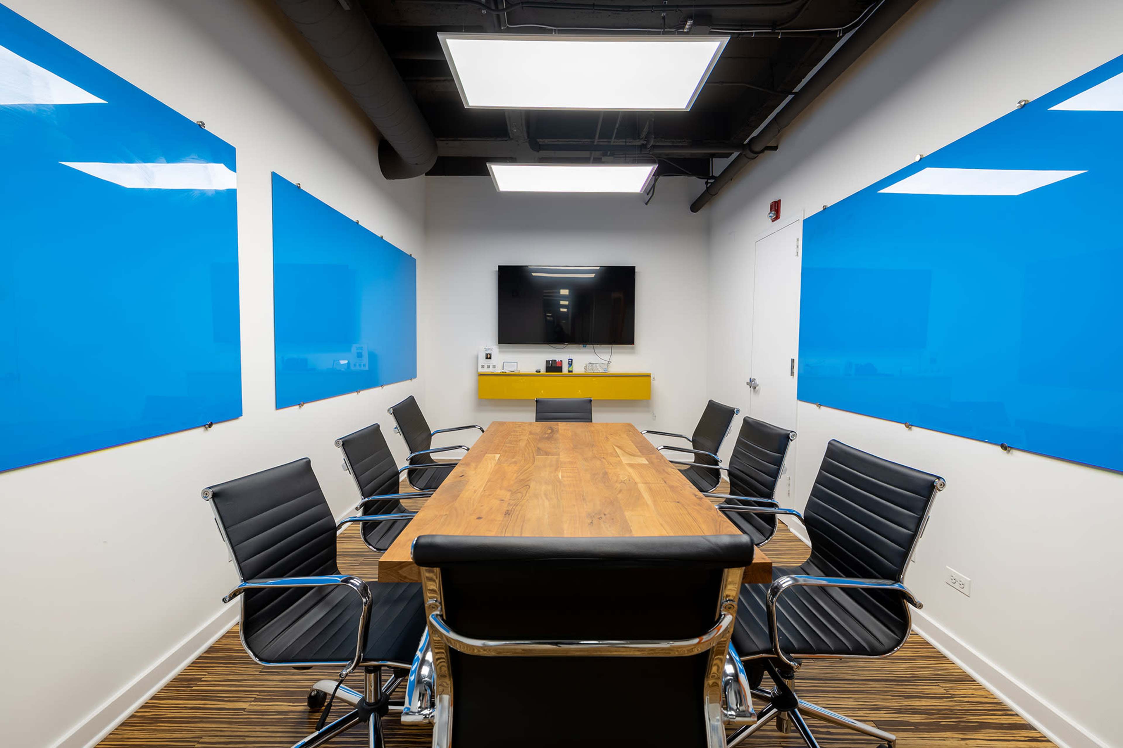 A modern conference room features a long wooden table surrounded by black chairs, with blue glass boards on the walls and a television mounted at one end.