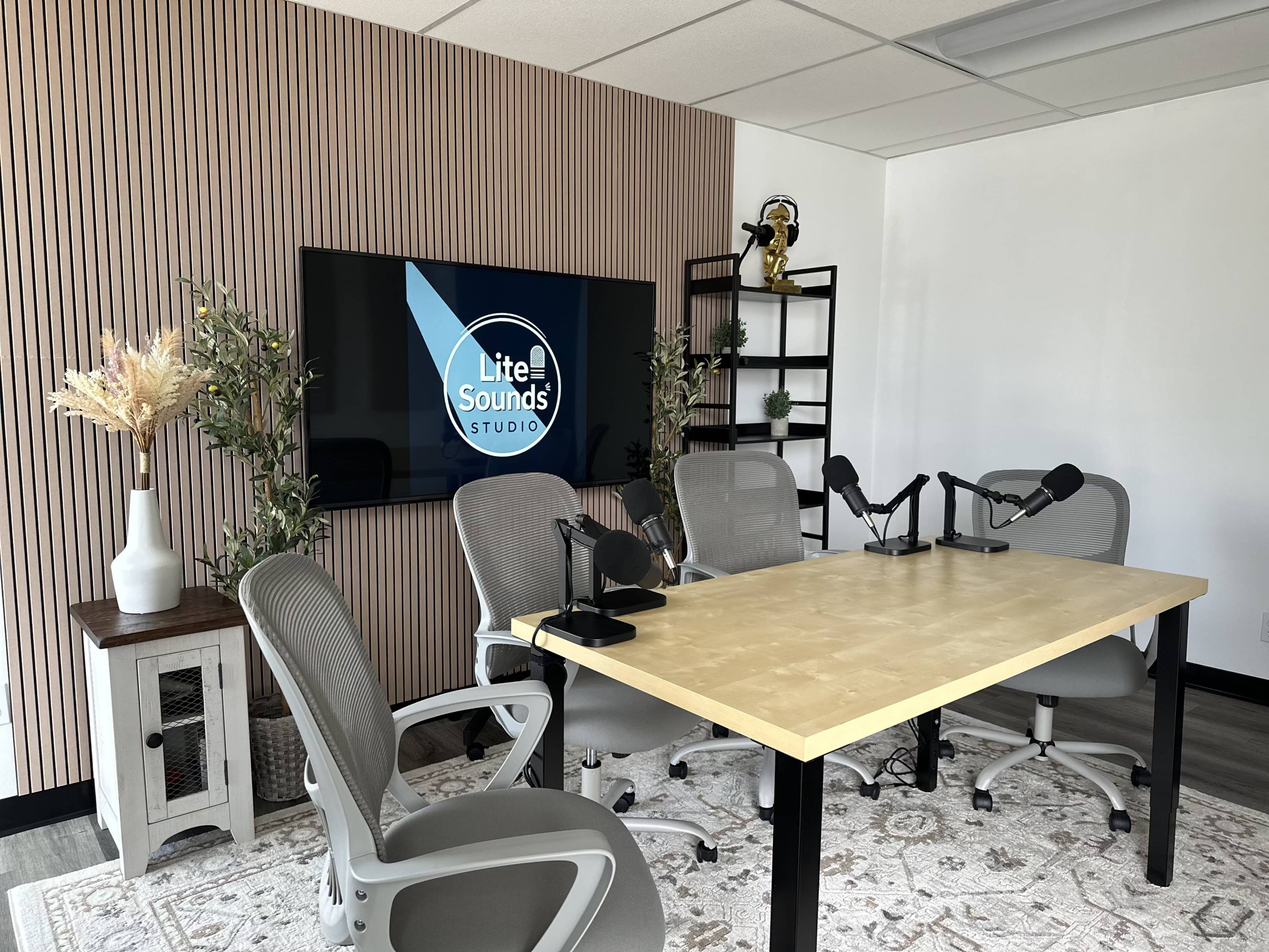 The image shows a well-organized studio space with a large screen displaying "Lite Sounds Studio," a table with microphones, and several chairs arranged around it.