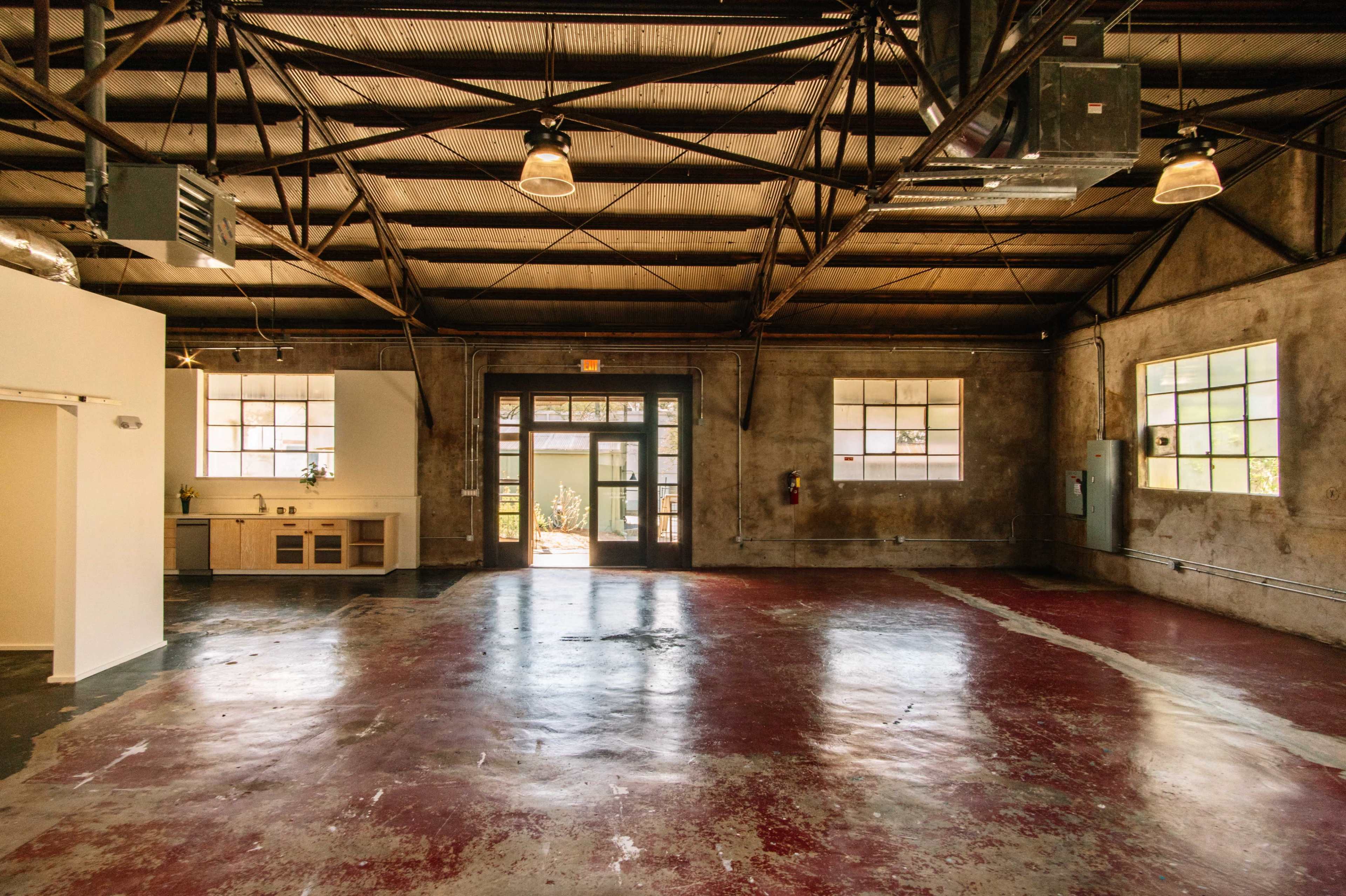 The image shows an empty industrial-style interior space with a polished concrete floor, large windows, and exposed metal beams in the ceiling.