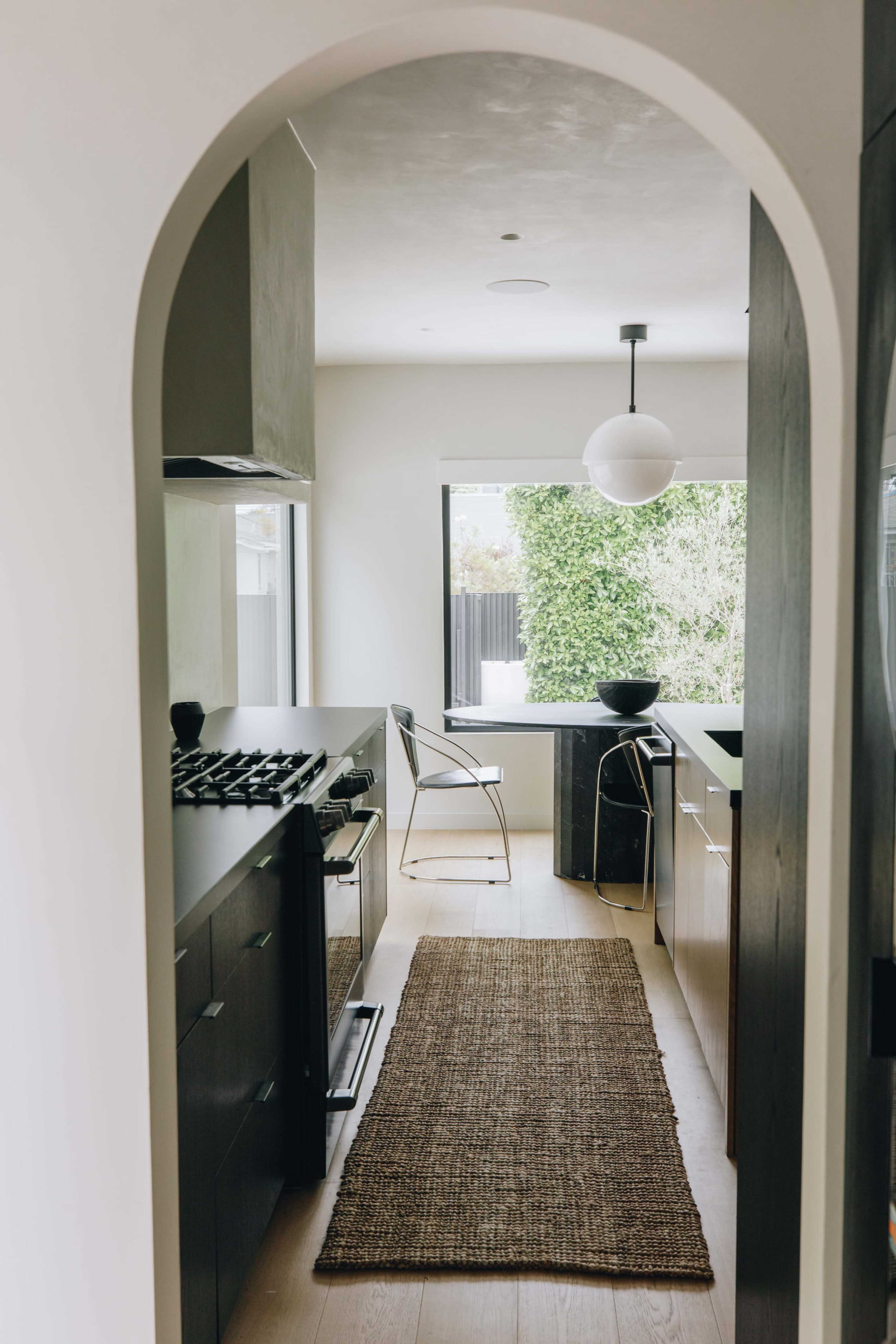 A view of a modern kitchen through an arched doorway, featuring a gas stove, minimalist cabinetry, and a window overlooking greenery.