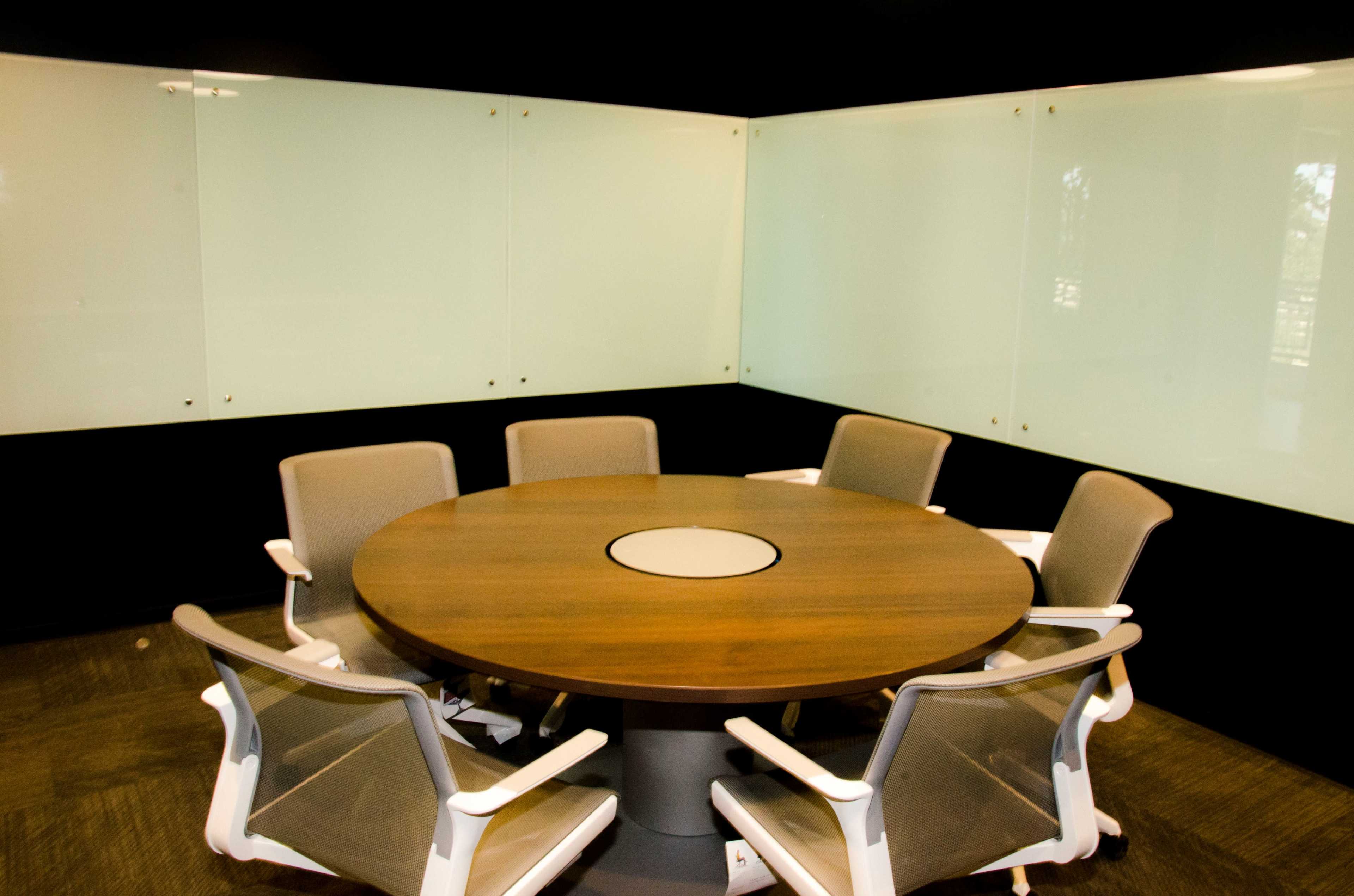 A modern conference room with a circular wooden table and six ergonomic chairs arranged around it, set against glass walls.
