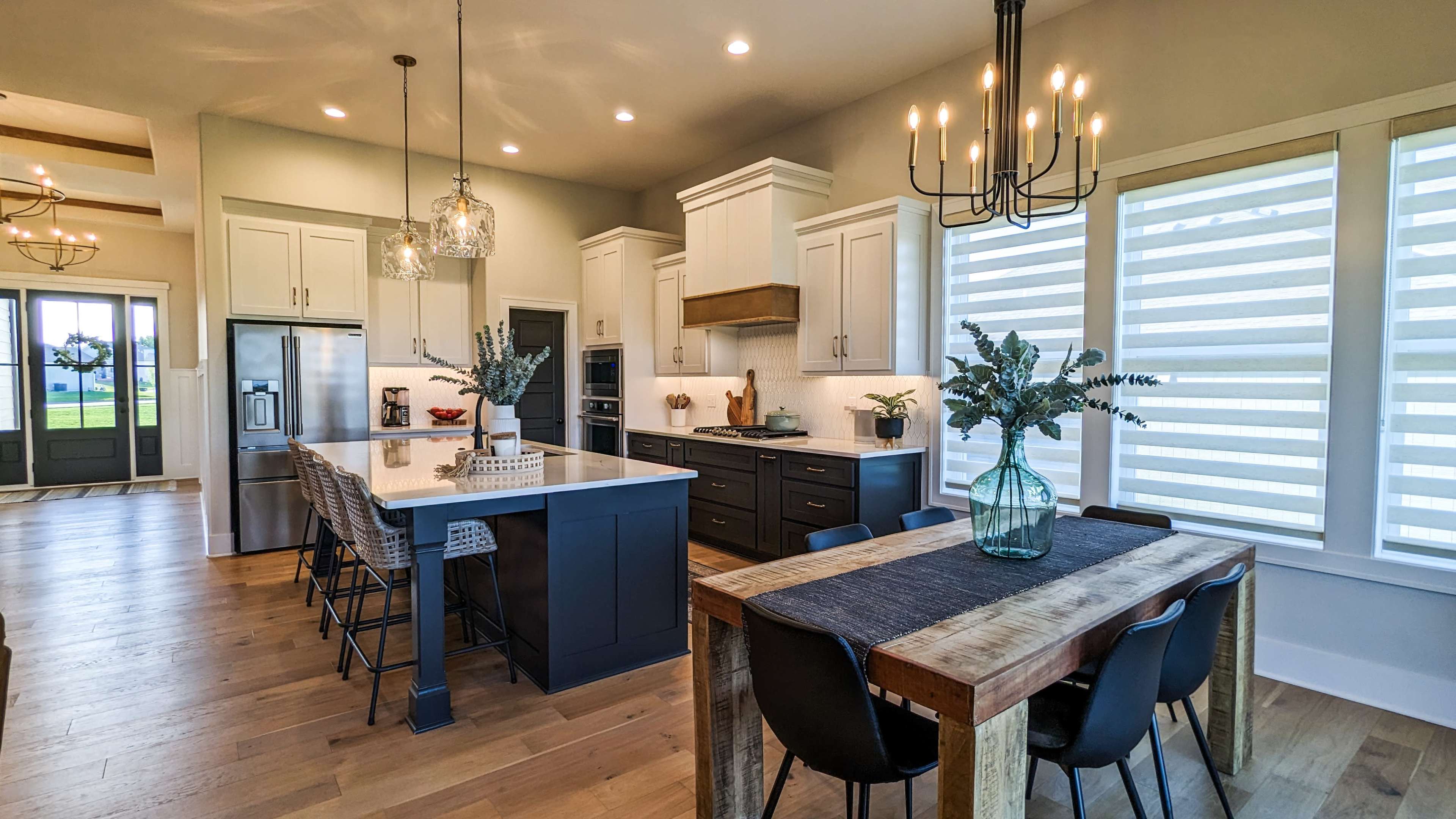 A modern kitchen with a large island, six bar stools, a dining table, and large windows with white window treatments.