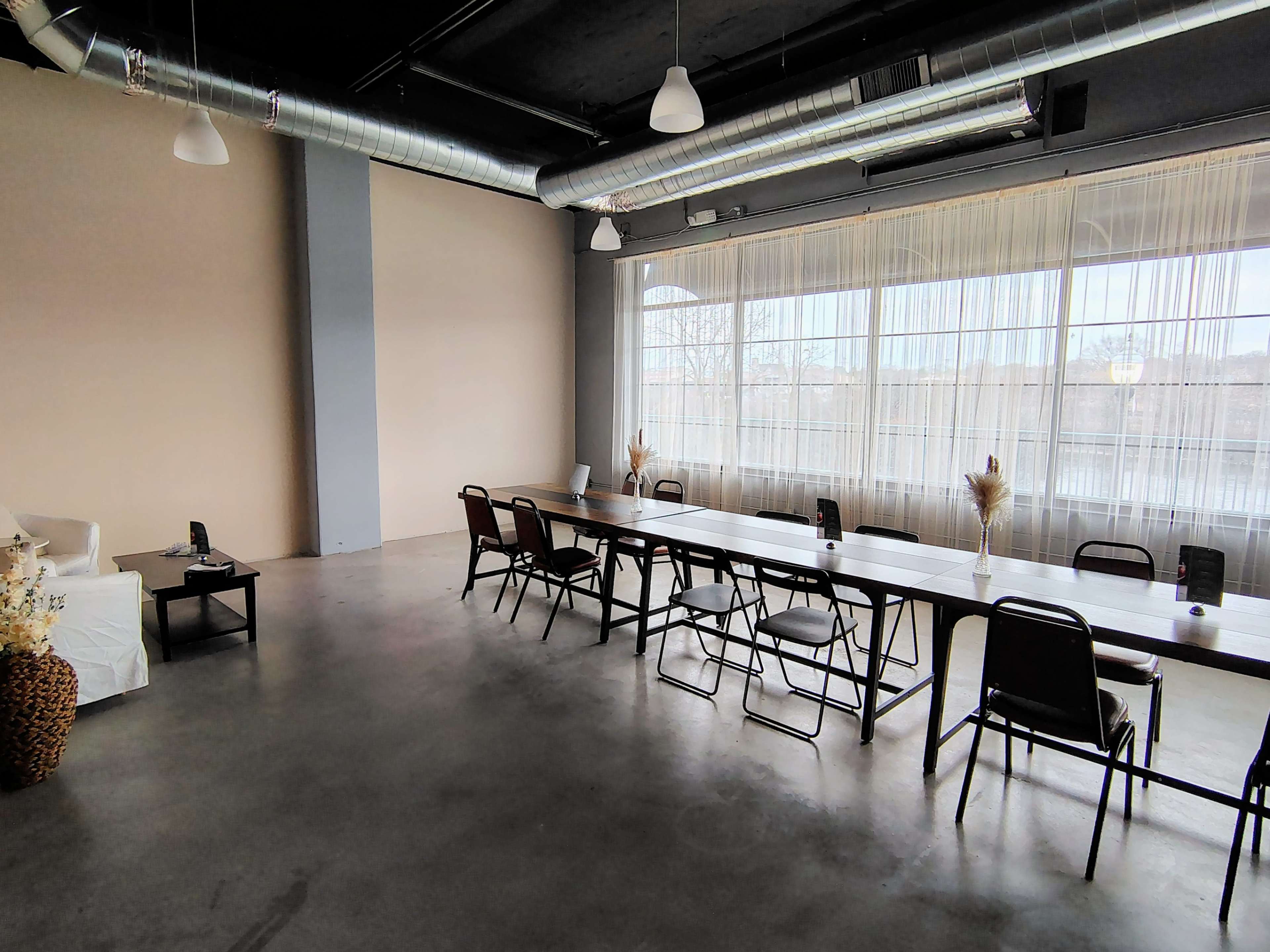 A spacious room features a long table surrounded by chairs, with large windows allowing natural light to fill the space.