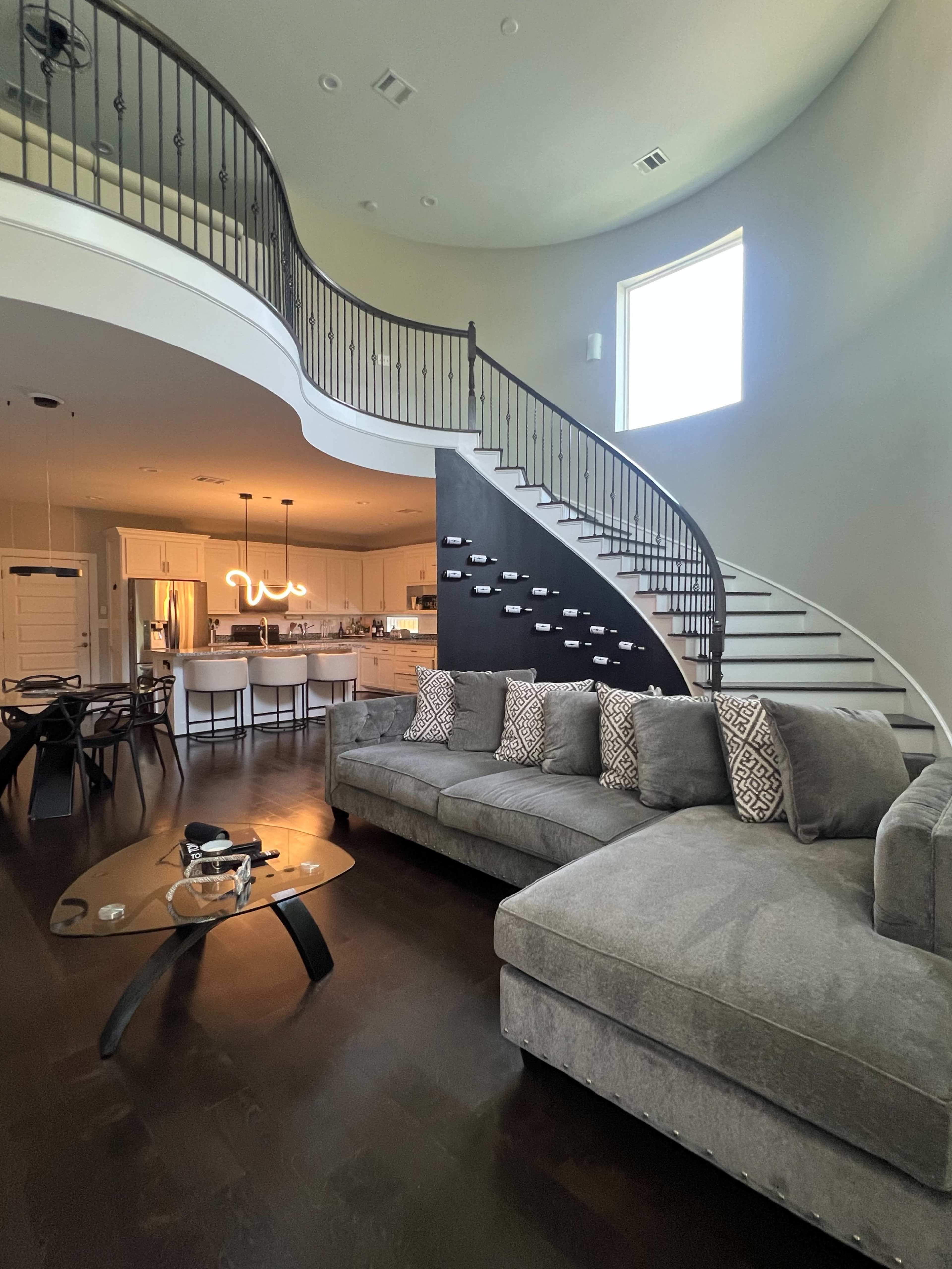 A modern living room with a large gray sectional sofa, a coffee table, and a sweeping staircase that leads to an upper floor.
