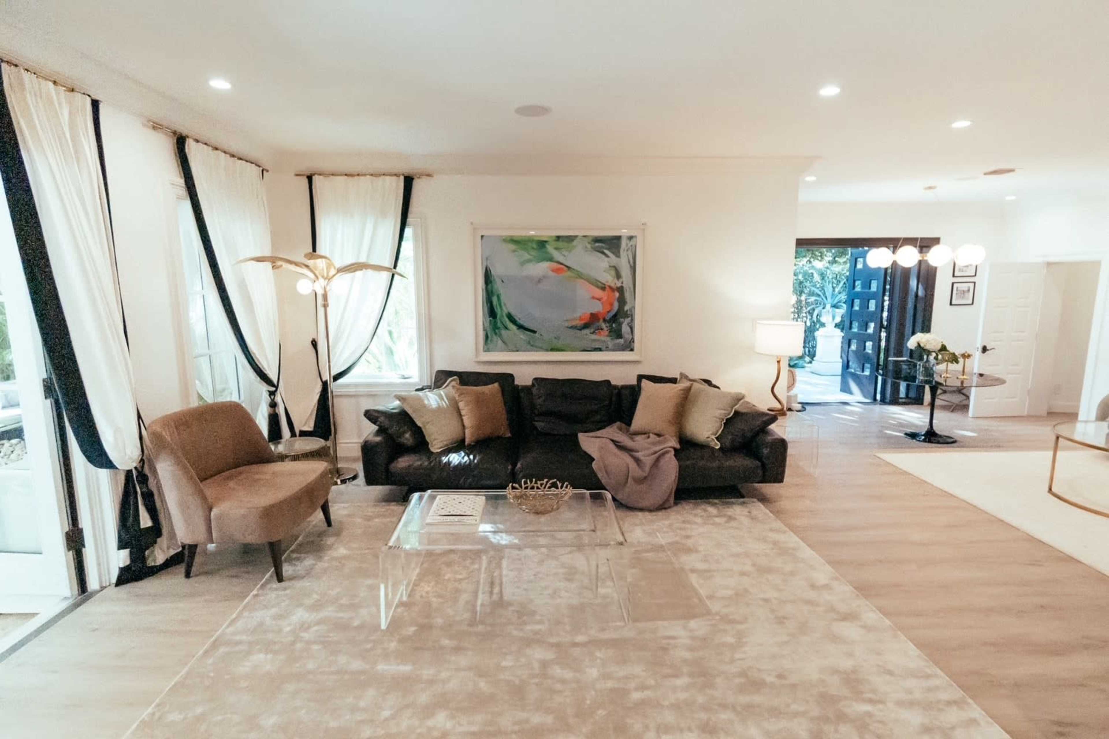 The image depicts a modern living room featuring a dark leather sofa, a large abstract painting on the wall, and light-colored flooring with a decorative rug.