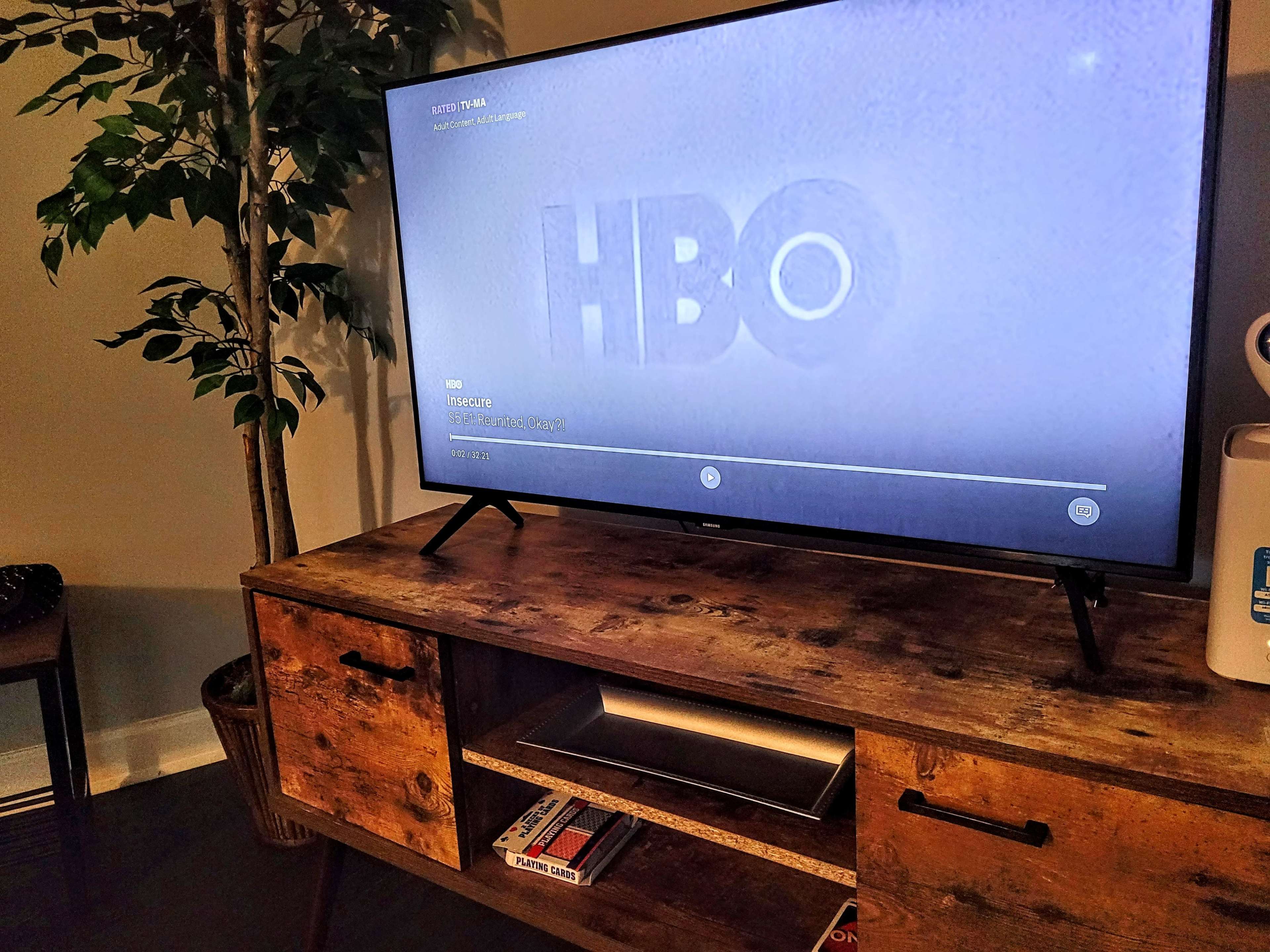A television displaying the HBO logo sits on a wooden media console, with a potted plant and a few items visible nearby.