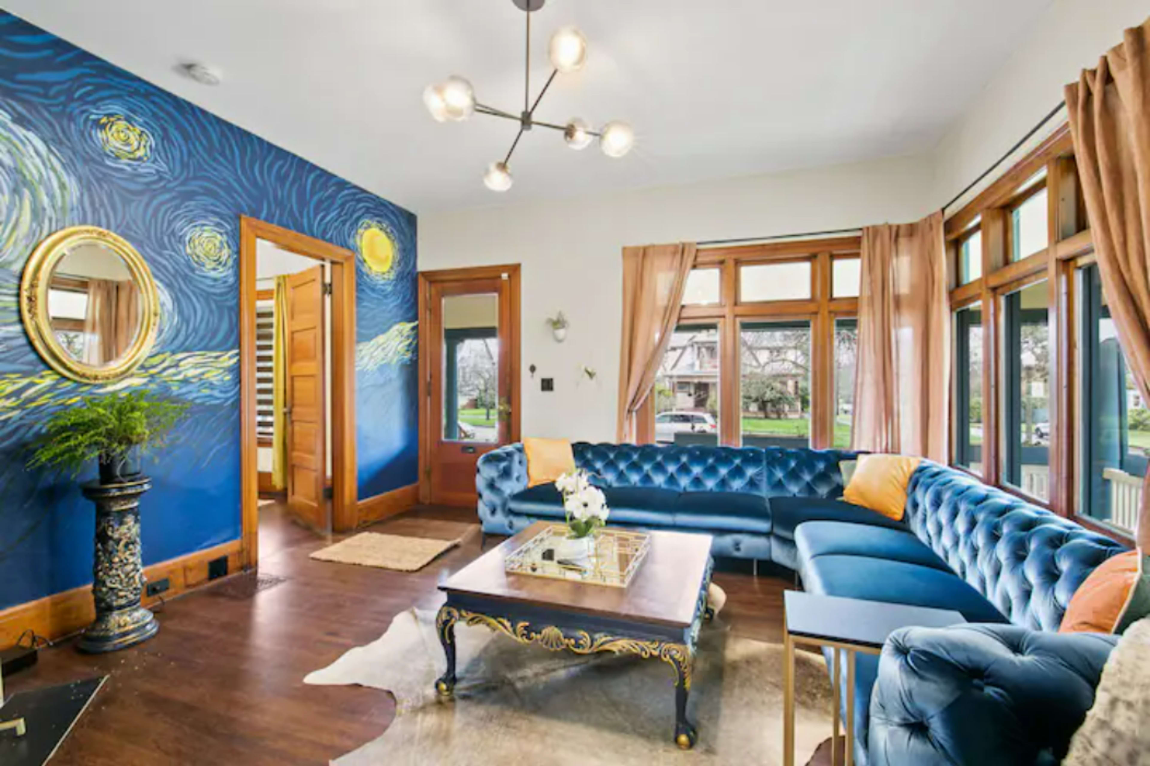 A living room features a blue velvet sectional sofa, a coffee table, and a mural of swirling stars on one wall, with large windows allowing natural light.