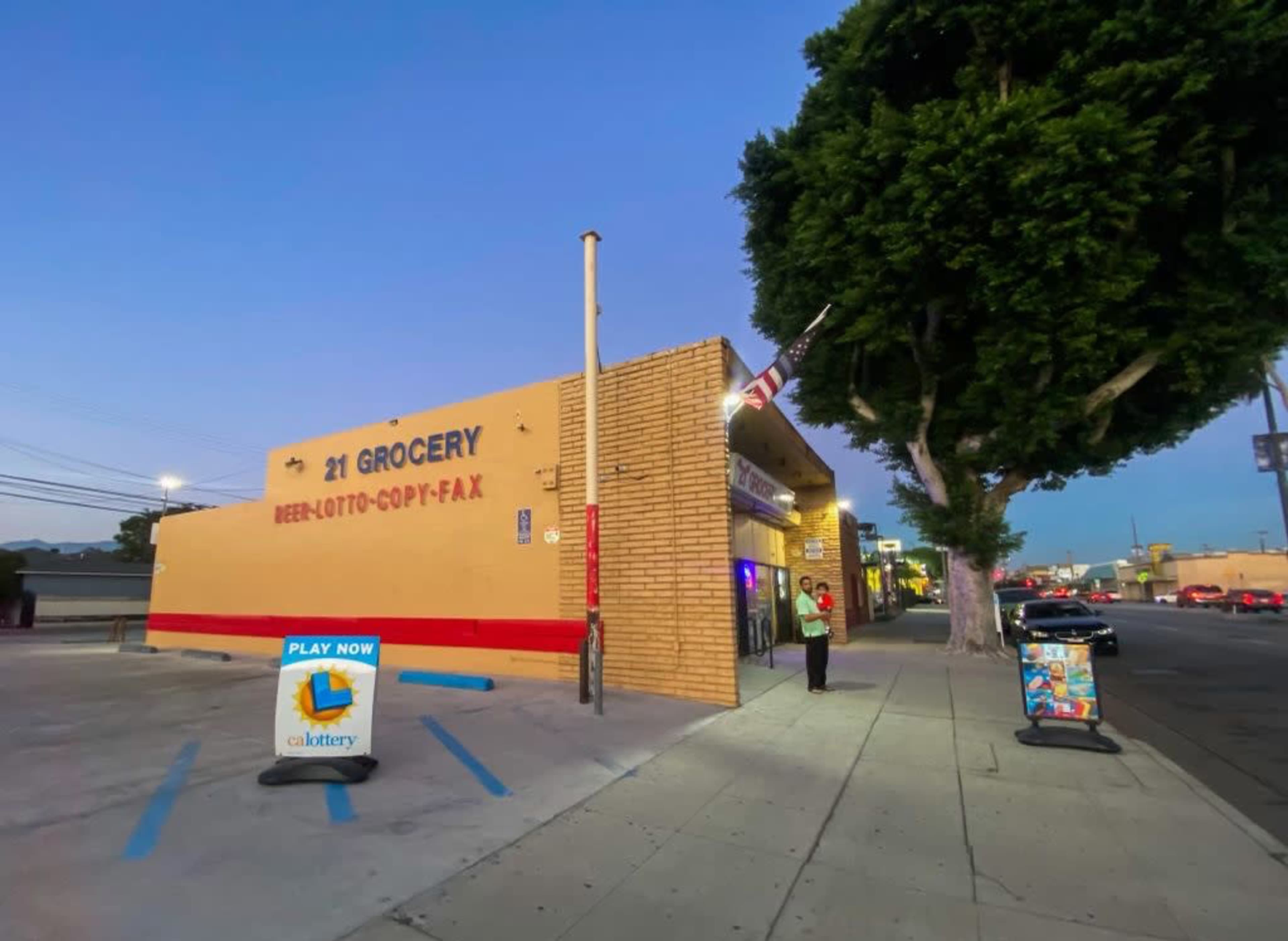 A grocery store with a sign displaying its services and a person standing outside on a sidewalk, at dusk.