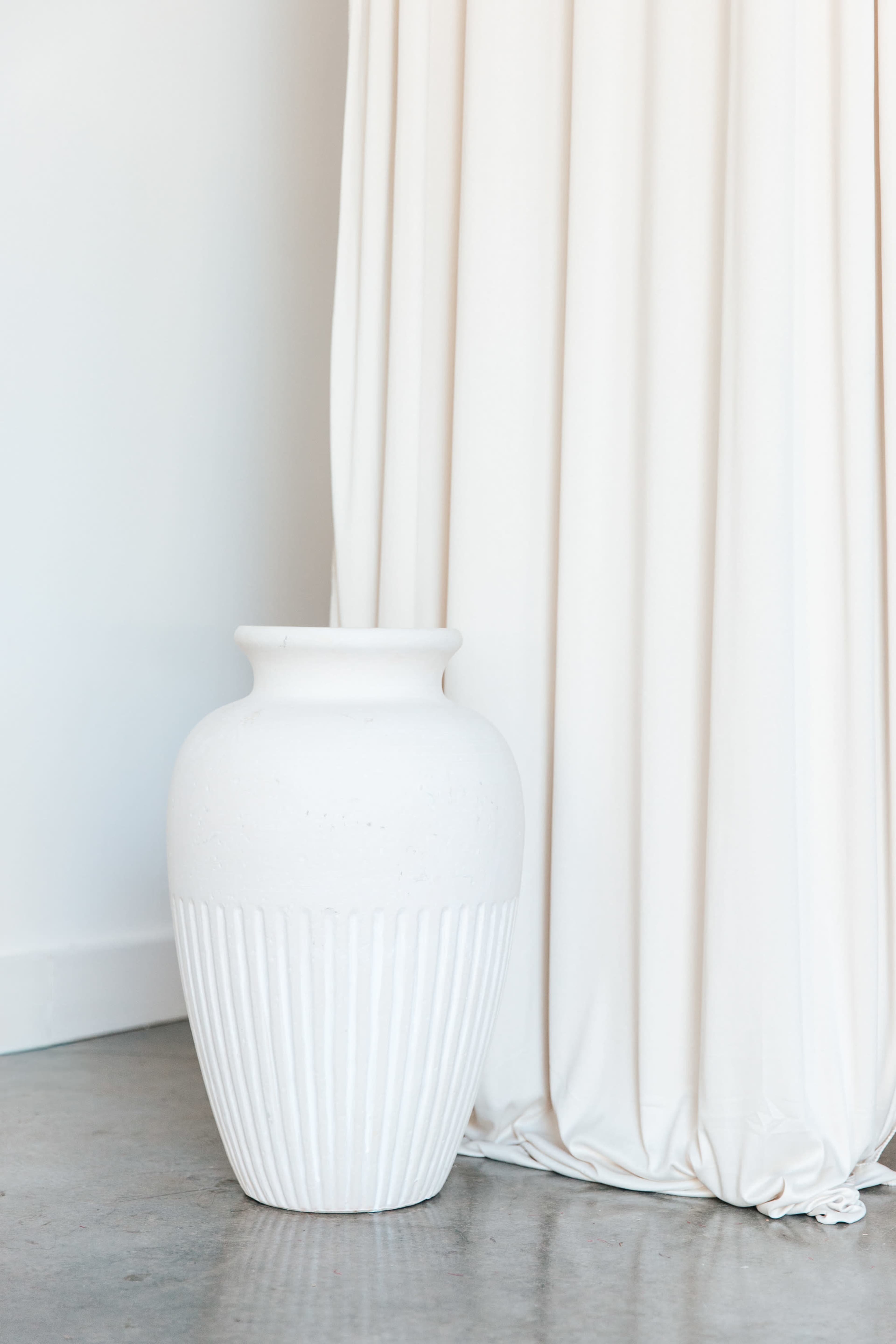 A white, textured ceramic vase stands next to a flowing white curtain on a concrete floor.