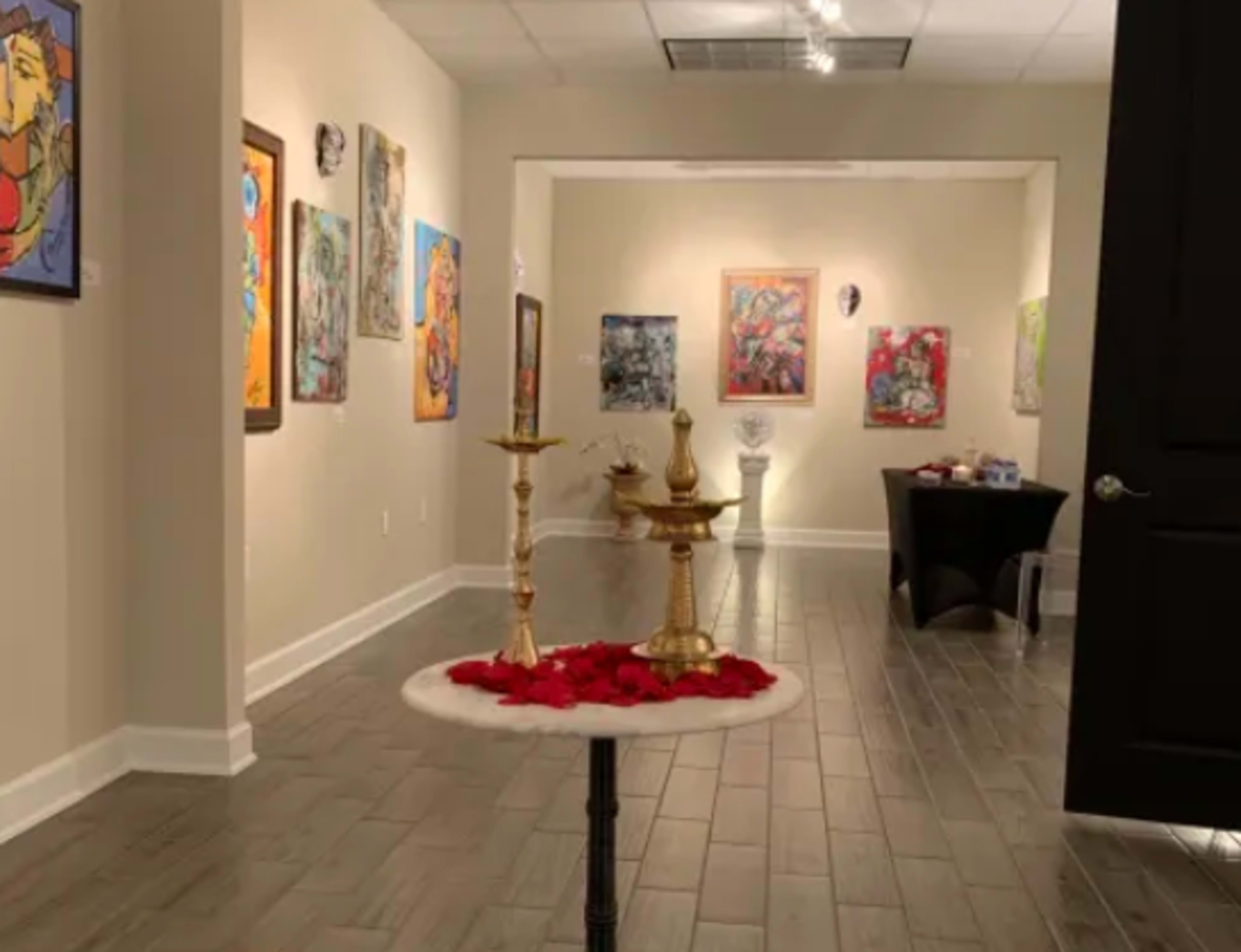 The image shows an art gallery adorned with colorful paintings on the walls, featuring a small table with decorative items and rose petals in the foreground.