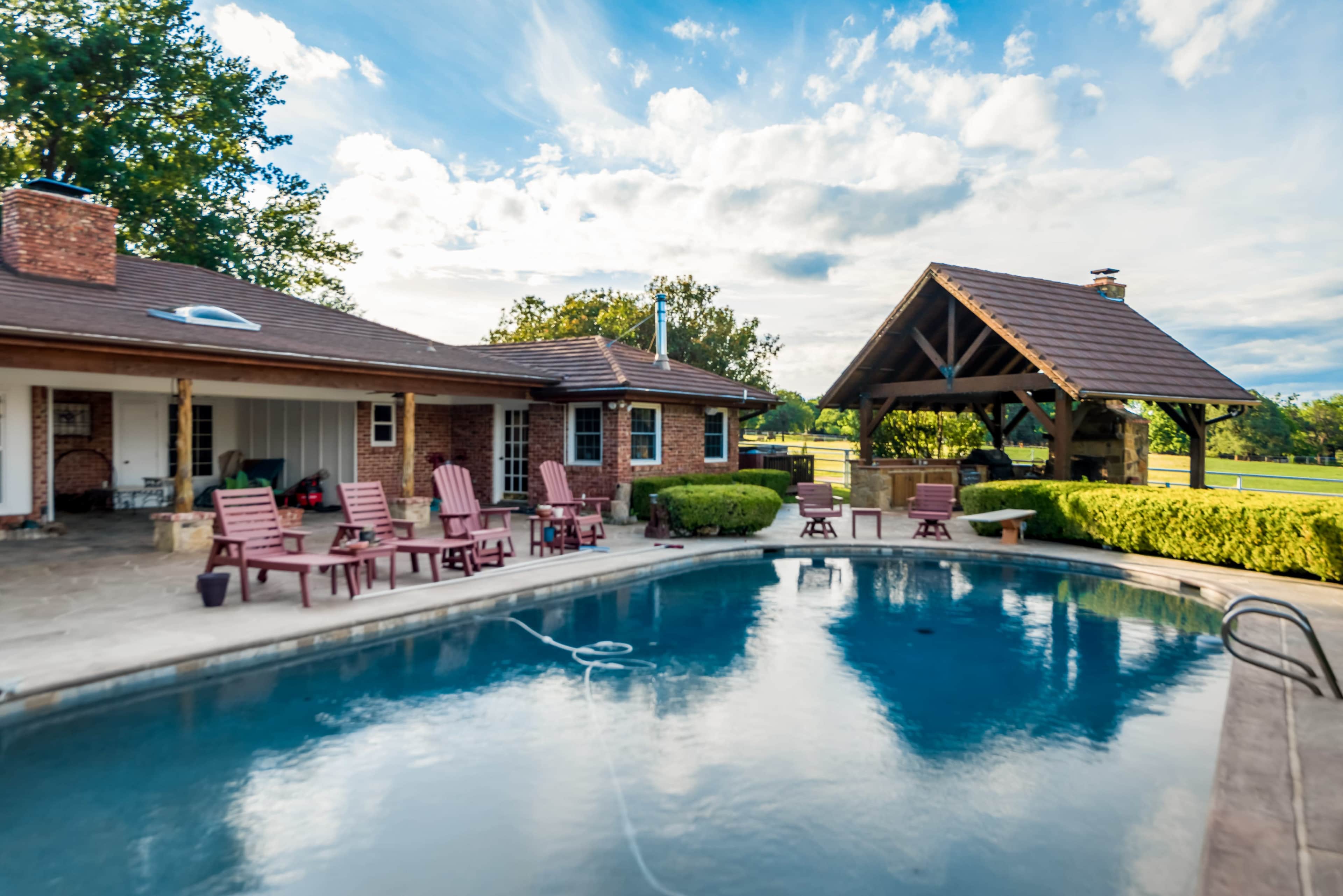 A backyard features a swimming pool surrounded by wooden lounge chairs and a pavilion with a sloped roof.