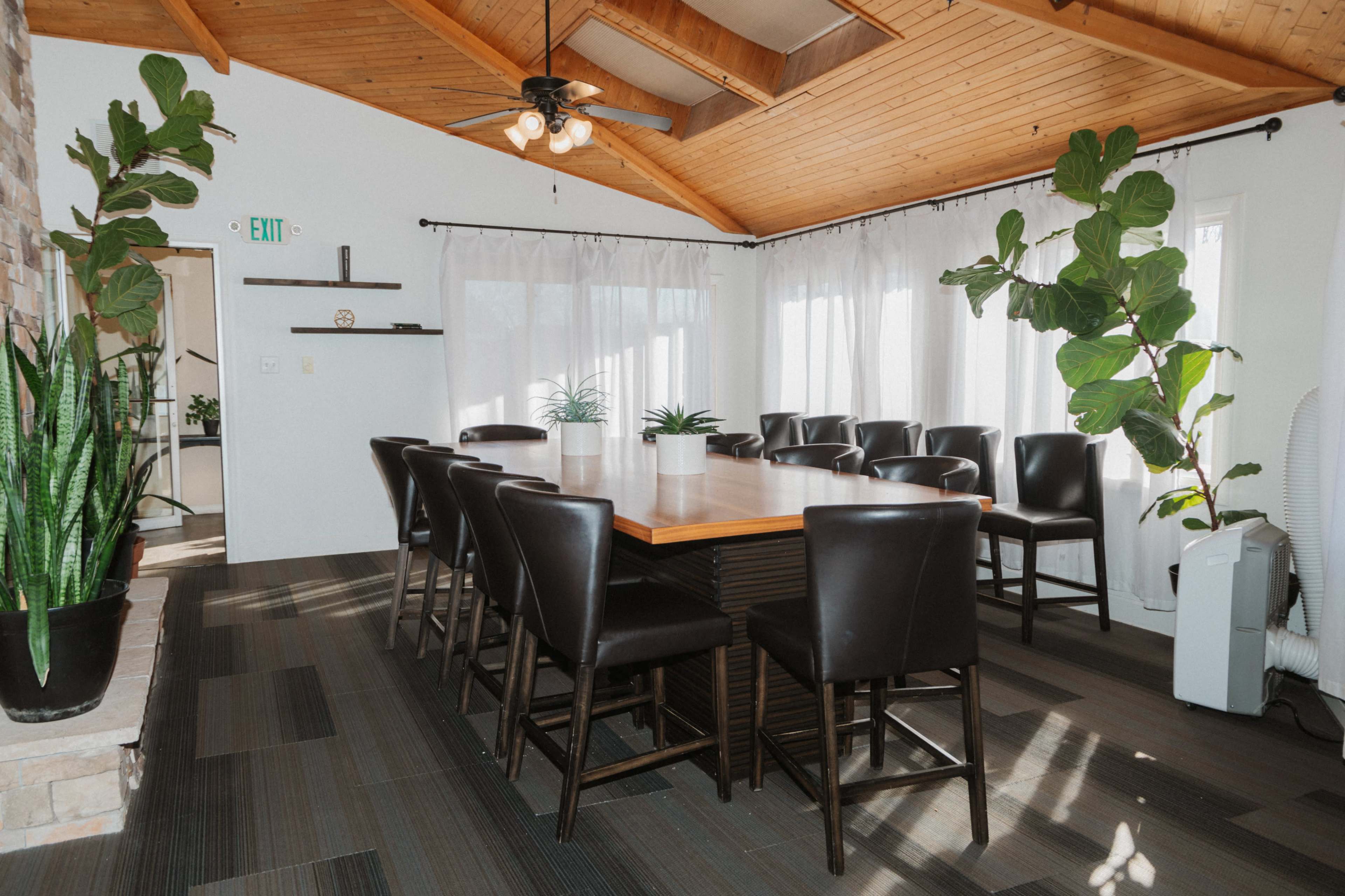 The image shows a conference room with a large wooden table surrounded by eight black chairs, featuring plants in the corners and large windows with sheer curtains.