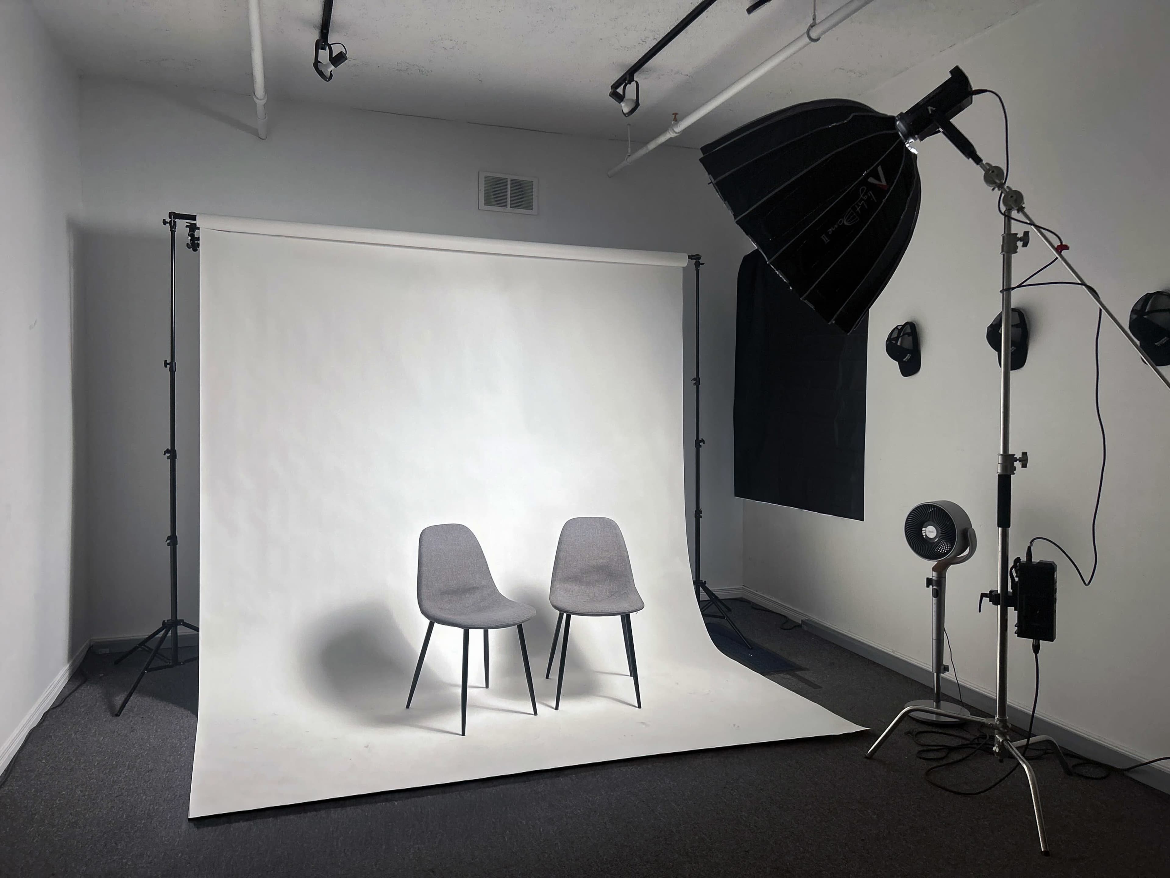 The image shows a photography studio setup with two gray chairs on a white backdrop, illuminated by a large softbox and a portable fan.